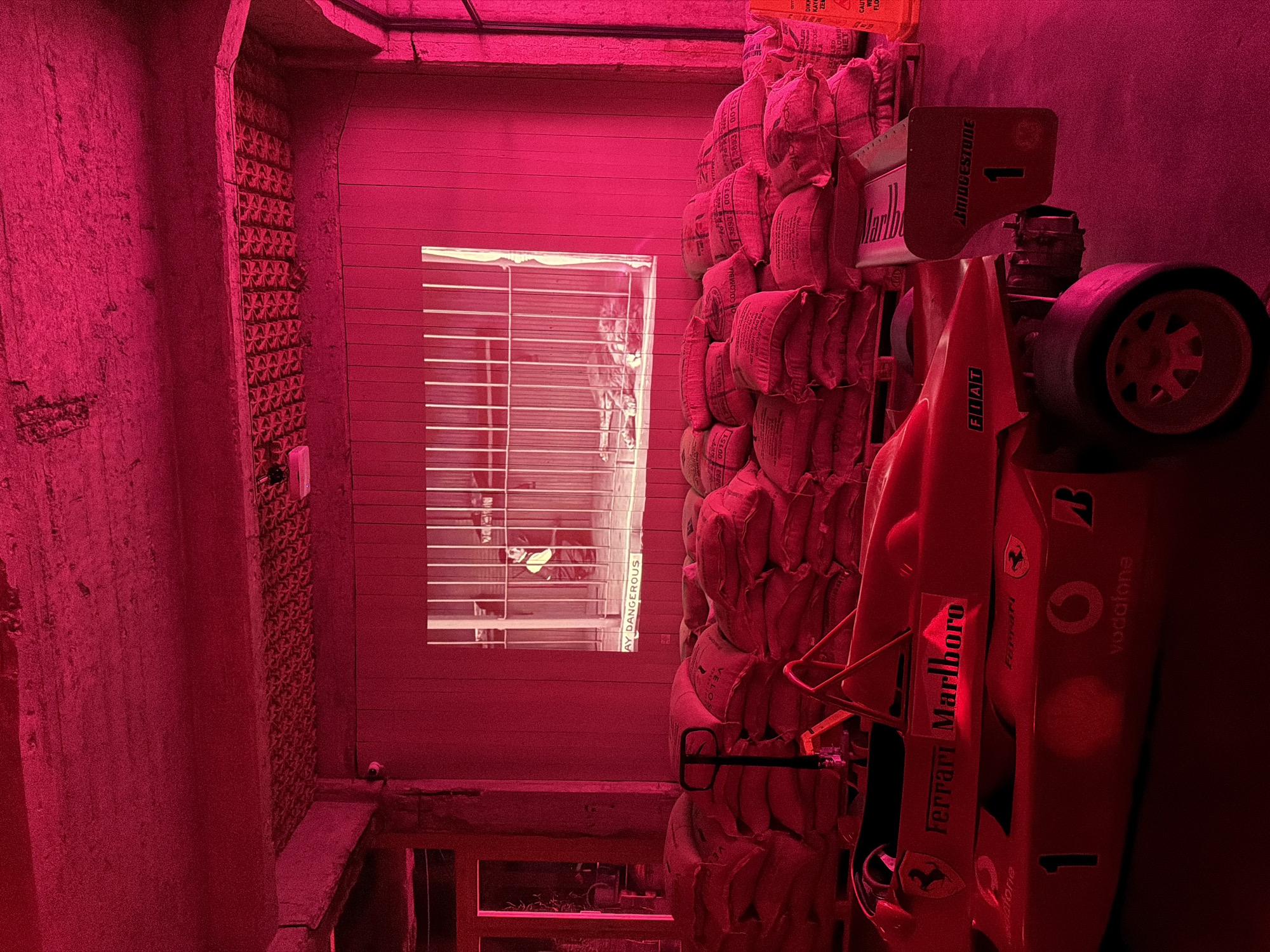 The image captures a dimly lit, indoor scene characterized by an intense red hue, likely from the ambient lighting. Stacks of burlap sacks fill a significant portion of the scene, and in front of these sacks is a red Ferrari Formula One race car replica with "Ferrari Marlboro" prominently displayed on the side. The car also features the Vodafone logo and Bridgestone on the rear wing. A partially visible window with horizontal blinds is present in the background. The overall setting appears to be a storage area, with concrete walls and a partially visible lower level or basement. The OCR data suggests the location is in Tbilisi, Georgia. The overall impression is a cluttered, somewhat chaotic scene with an unusual color cast.