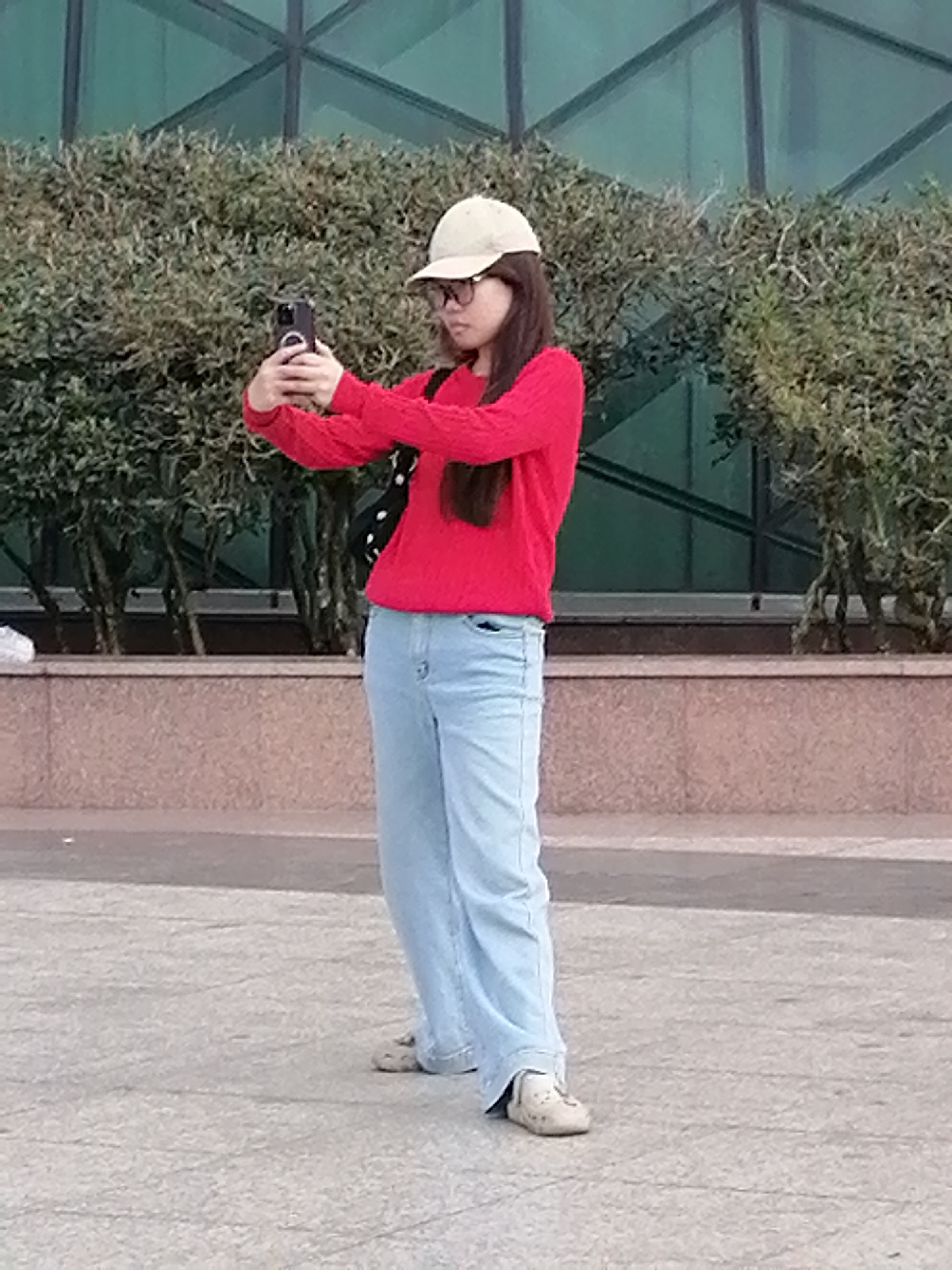 A young woman stands outdoors, holding a smartphone to take a selfie. She is wearing a red long-sleeved sweater, light blue wide-leg jeans, a light-colored baseball cap, and glasses. She has dark hair that falls past her shoulders. The background features dense green shrubbery and a building with a green glass facade. The ground is paved with a textured gray material. The scene appears to be a public space, possibly a park or plaza. There is no indication this image was taken in Da Lat, Vietnam.