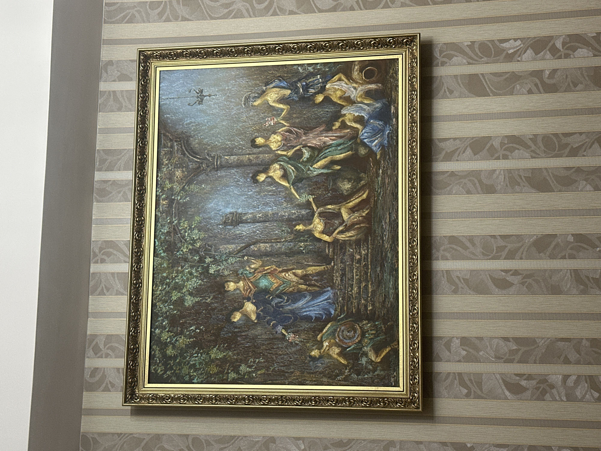 The image shows a framed painting hanging on a wallpapered wall. The wallpaper has horizontal stripes with a floral pattern. The painting depicts a scene with multiple figures in a natural setting. The figures appear to be stylized and possibly mythological. The painting has a gold ornate frame. The wall is located next to a white wall, suggesting it is indoors. The location is not discernible in the image, so the city of Yerevan, Armenia, cannot be confirmed based on the image alone.