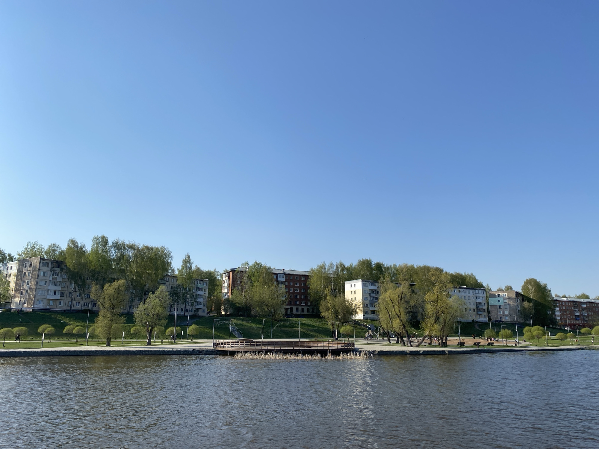 The image depicts a waterfront scene in Votkinsk, Russia, likely during a spring day given the trees displaying fresh green foliage. In the foreground, there is a body of water with gentle ripples across its surface. A wooden platform or pier extends slightly into the water. The shoreline is lined with a paved walkway and dotted with trees. Beyond the walkway, there is a grassy incline leading up to a row of buildings. The buildings are multi-story, residential structures, typical of Soviet-era architecture. The sky is clear and blue, suggesting fair weather. There are people walking or sitting along the promenade.