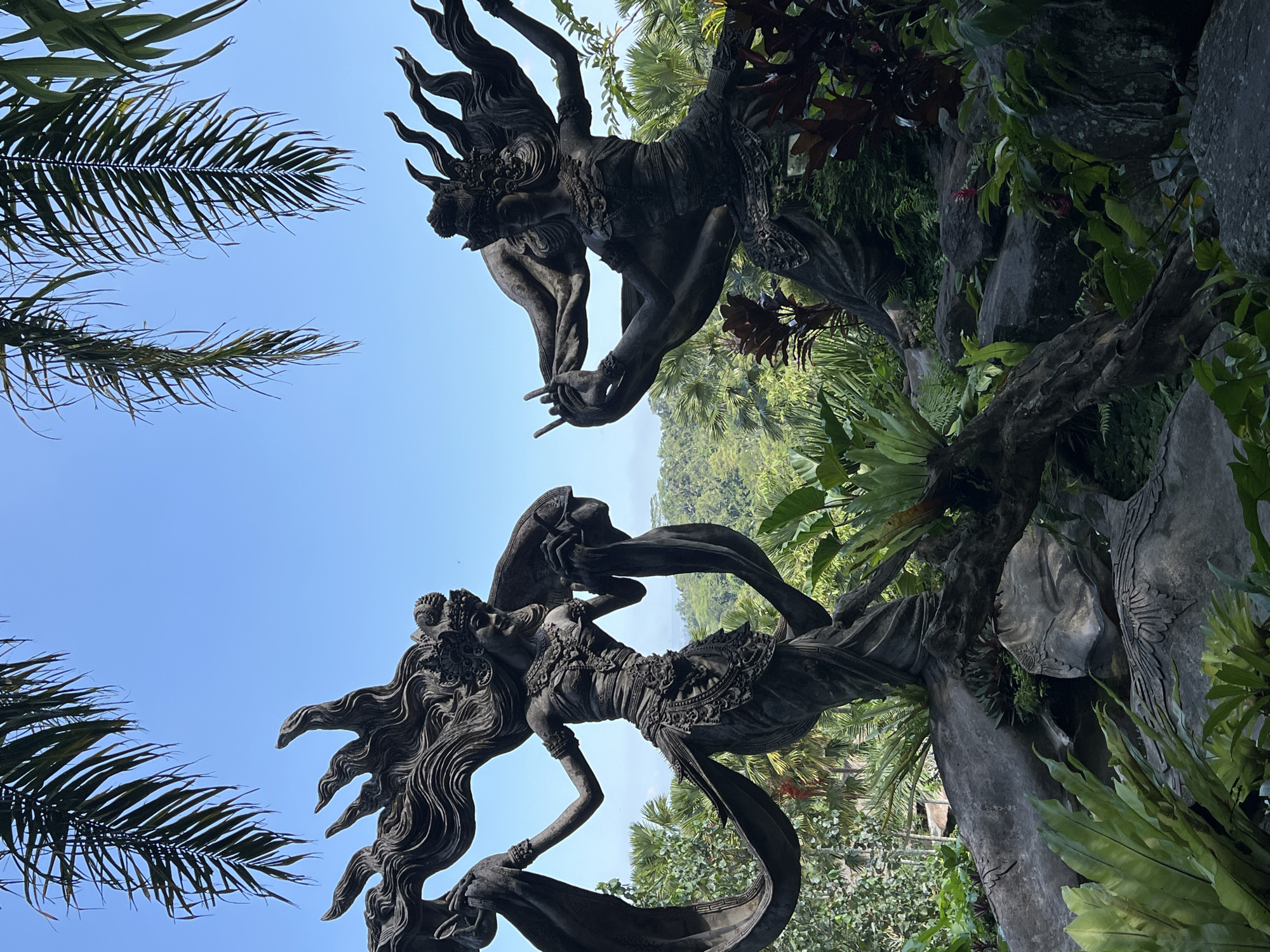 The image shows two Balinese statues amidst lush greenery and a clear blue sky. The statues are made of dark stone or metal. The lower statue is a female figure with long flowing hair, adorned with intricate jewelry and flowing garments. The upper statue is a male figure with a fiery headdress and expressive gestures. They appear to be reaching for each other. Palm fronds frame the left side of the image. The background reveals a landscape of rolling hills covered in dense vegetation. The location is Kedewatan, Indonesia.