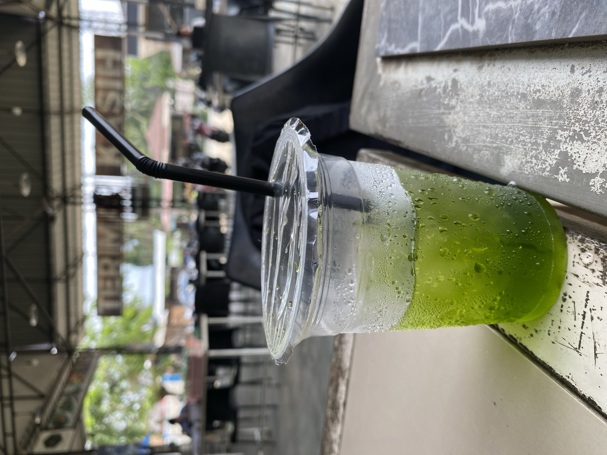 The image captures a green beverage in a clear plastic cup with a black straw, placed on a weathered, gray surface. Condensation droplets cling to the cup's exterior. The background reveals an outdoor setting, possibly a cafe or open-air establishment in Seuneubok Teungoh, Indonesia, as indicated by the urban environment with metal structures, buildings, and blurred figures and vehicles. The image is slightly rotated.