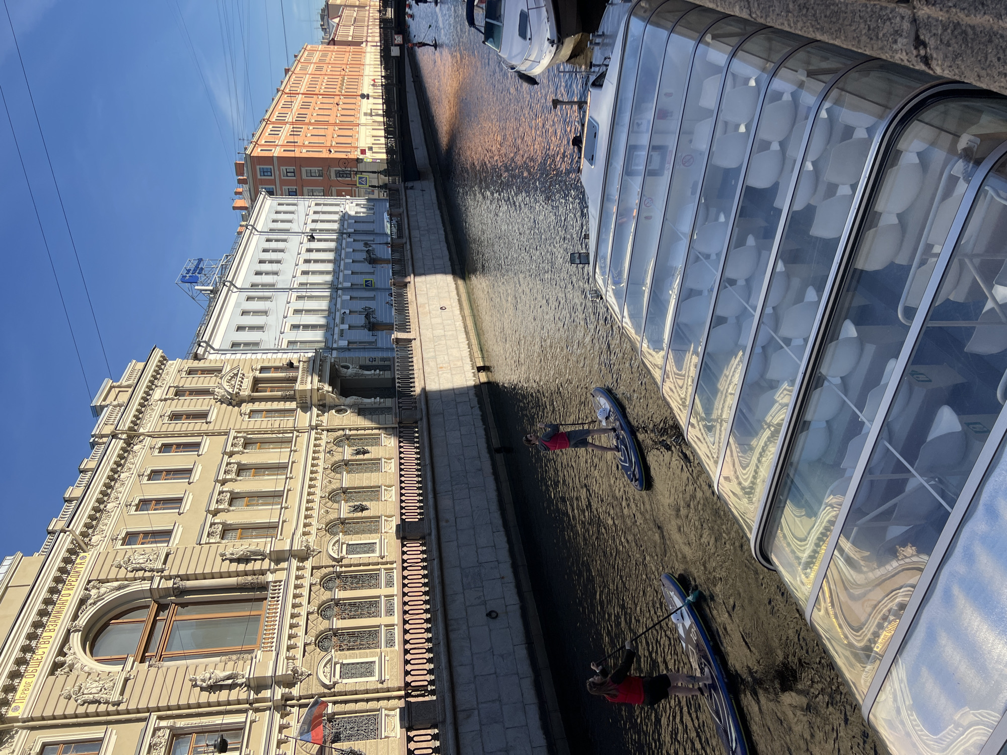 This is an eye-level, medium shot of a canal in Saint Petersburg, Russia, with a boat in the foreground and buildings lining the canal. The canal is dark and reflects the sunlight, creating a shimmering effect. Two individuals are paddleboarding on the canal, each on their own board. The person in the foreground is closer to the camera and is on the left side of the canal. They are wearing a red vest and dark shorts and are holding a paddle. The second person is further down the canal, on the right side, also wearing a red vest and dark shorts, and holding a paddle.
In the foreground, a boat with a glass roof is moored to the canal's edge. The boat has rows of white seats visible through the transparent roof. Behind the paddleboarders, a white motorboat is docked.
The canal is bordered by a stone embankment. Across the canal, a row of historic buildings stands, featuring ornate facades with intricate carvings and numerous windows. The buildings vary in color, from light beige to pale blue and orange. The sky is clear and blue, suggesting a sunny day. The image captures a serene urban scene with a blend of recreational activity and architectural grandeur.