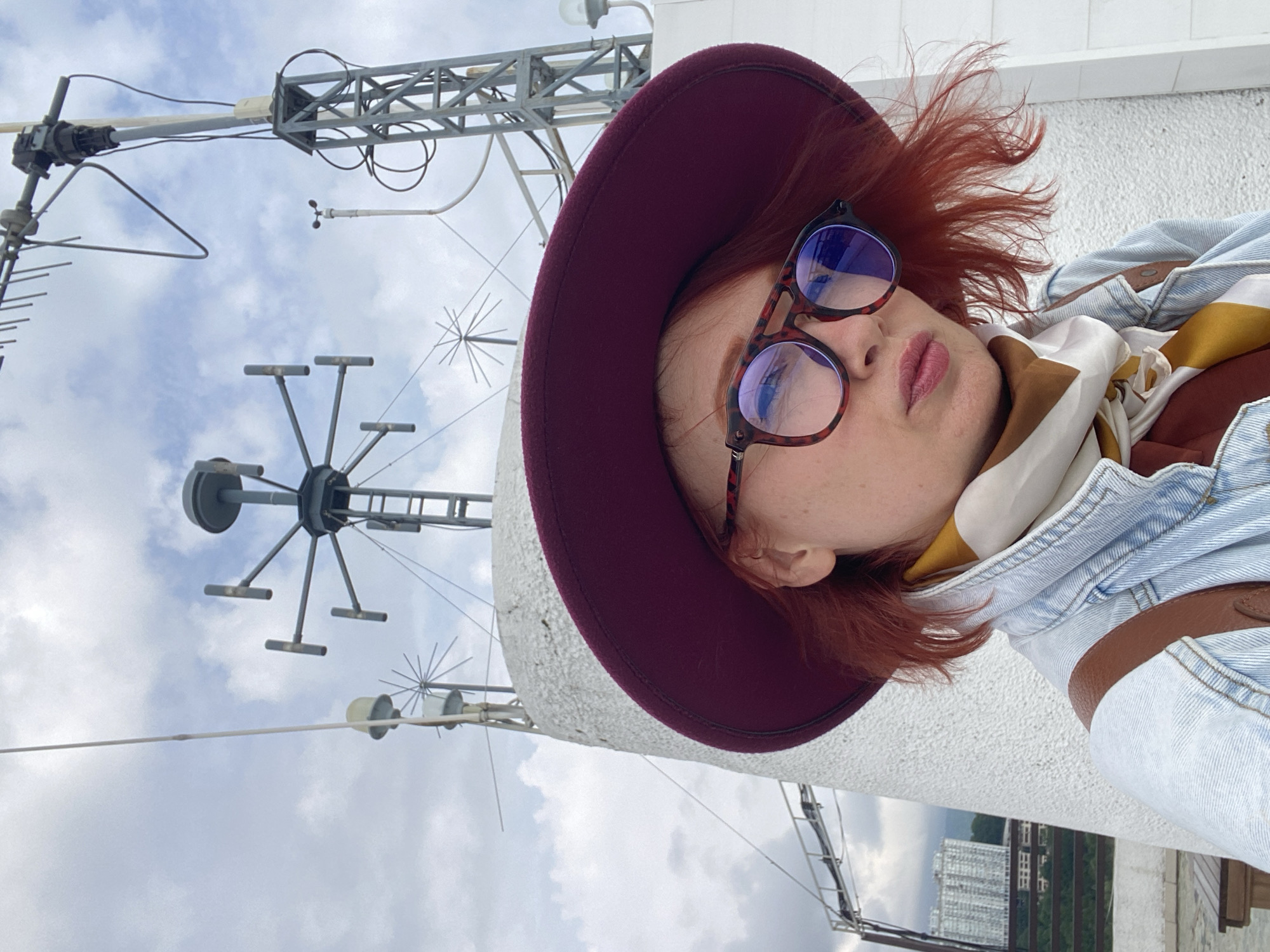 A woman wearing a maroon hat and glasses takes a selfie on what appears to be the roof of a building in Sochi, Russia. A white scarf with a yellow and brown pattern is visible around her neck, as well as a denim jacket. Behind her are several metal structures that resemble antennas or weather monitoring equipment. In the background, buildings, trees, and cloudy skies can be seen.