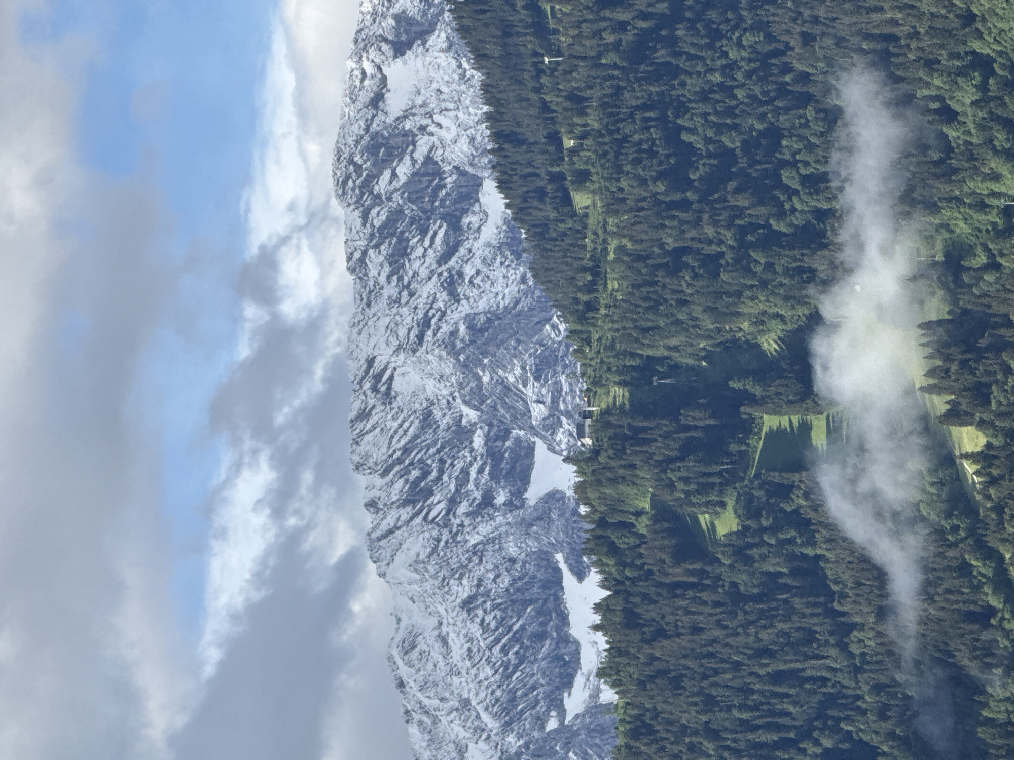 This image captures a view of the Zugspitze massif, the highest peak in Germany, located near Garmisch-Partenkirchen. The mountain range is partially covered in snow, contrasting with the dense, dark green coniferous forest that blankets the lower slopes. A winding green path, likely a hiking trail or ski slope, is visible cutting through the forest. Low-lying clouds or mist drift through the lower portion of the forest. In the distance, on a higher part of the forested slope, a small cluster of buildings with white roofs can be seen. The sky above is a mix of blue patches and grey clouds. There are no people visible in the image.