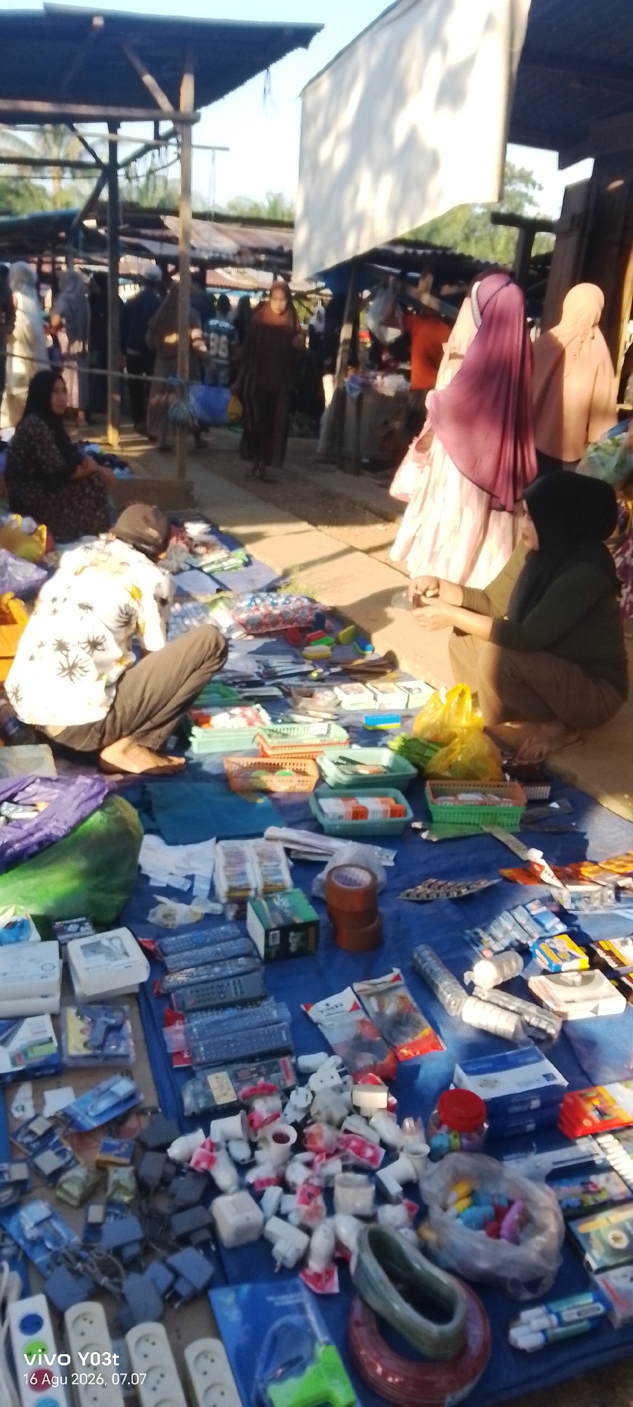 The image depicts an outdoor market scene in Palosilae, Indonesia, likely during daylight hours. In the foreground, a blue tarp is spread out on the ground, displaying a variety of goods for sale. These include numerous remote controls, electronic adapters, power strips, small white LED lights, and what appear to be small packages of batteries or other electronic components. There are also some packaged goods with colorful designs, possibly snacks or small toys. Two individuals are visible in the foreground: one person, partially obscured, is crouched down examining the items on the tarp. Another person, wearing a dark hijab and olive-colored clothing, is also crouched, appearing to be engaged in a transaction or organizing goods.
In the mid-ground, several people are walking and browsing. Some individuals are wearing hijabs, indicating a cultural context. Structures made of wood and corrugated metal, typical of market stalls, are present. A large white tarp is stretched overhead, possibly to provide shade. In the background, more market stalls and people are visible, with trees and foliage suggesting a rural or semi-urban setting. The overall impression is of a busy, local marketplace where goods are displayed on the ground for sale. The lighting suggests it could be morning or late afternoon.