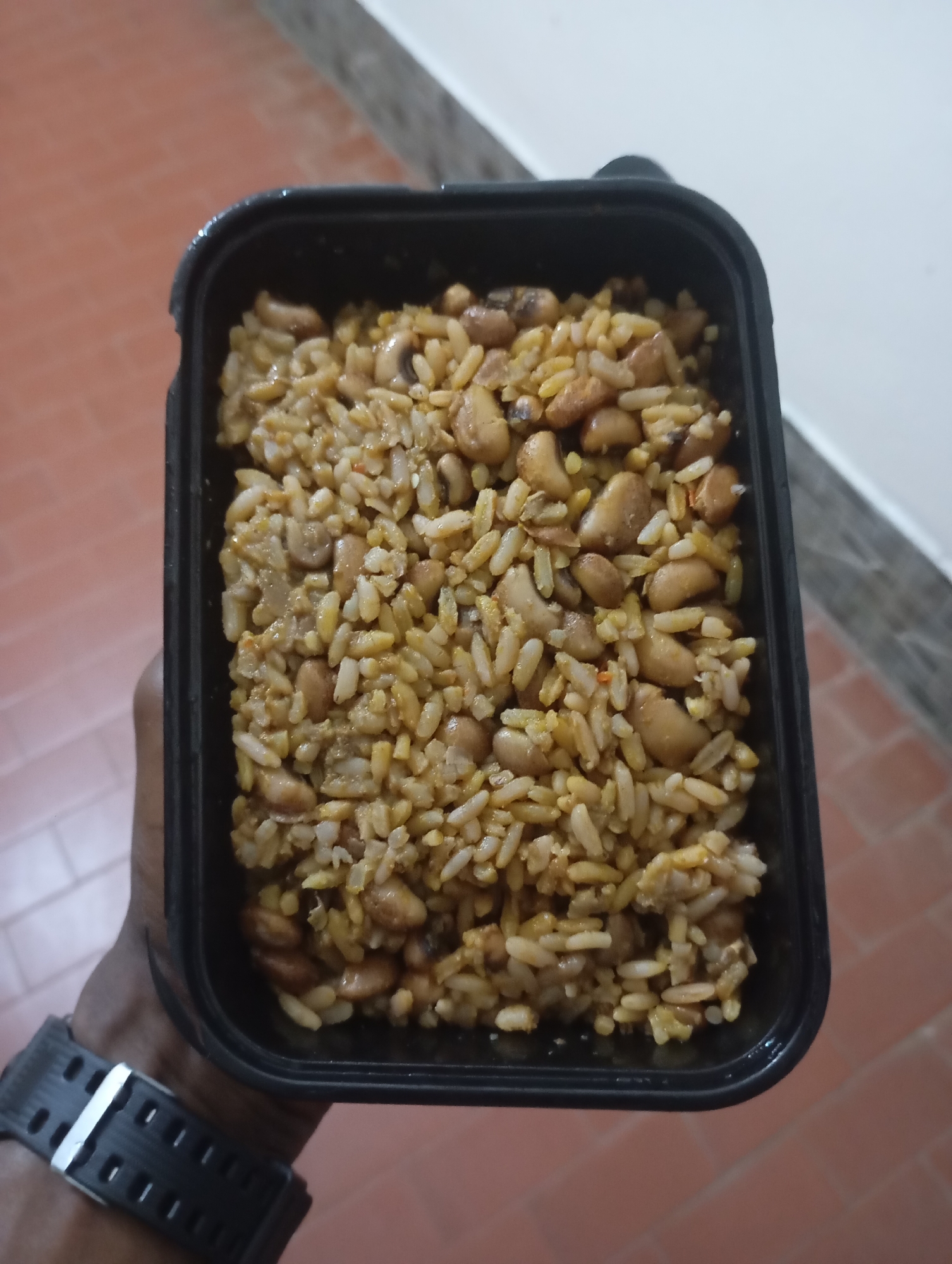 A top-down, close-up shot shows a black plastic container filled with rice and beans, held by a person's hand. The container is positioned in the lower center of the frame. The rice appears to be cooked and mixed with beans, giving it a yellowish-brown hue. The person's forearm and wrist are visible on the left side, wearing a black wristwatch with a metal clasp. The background consists of a reddish-brown tiled floor and a white wall. The image was taken in Jos, Nigeria.
