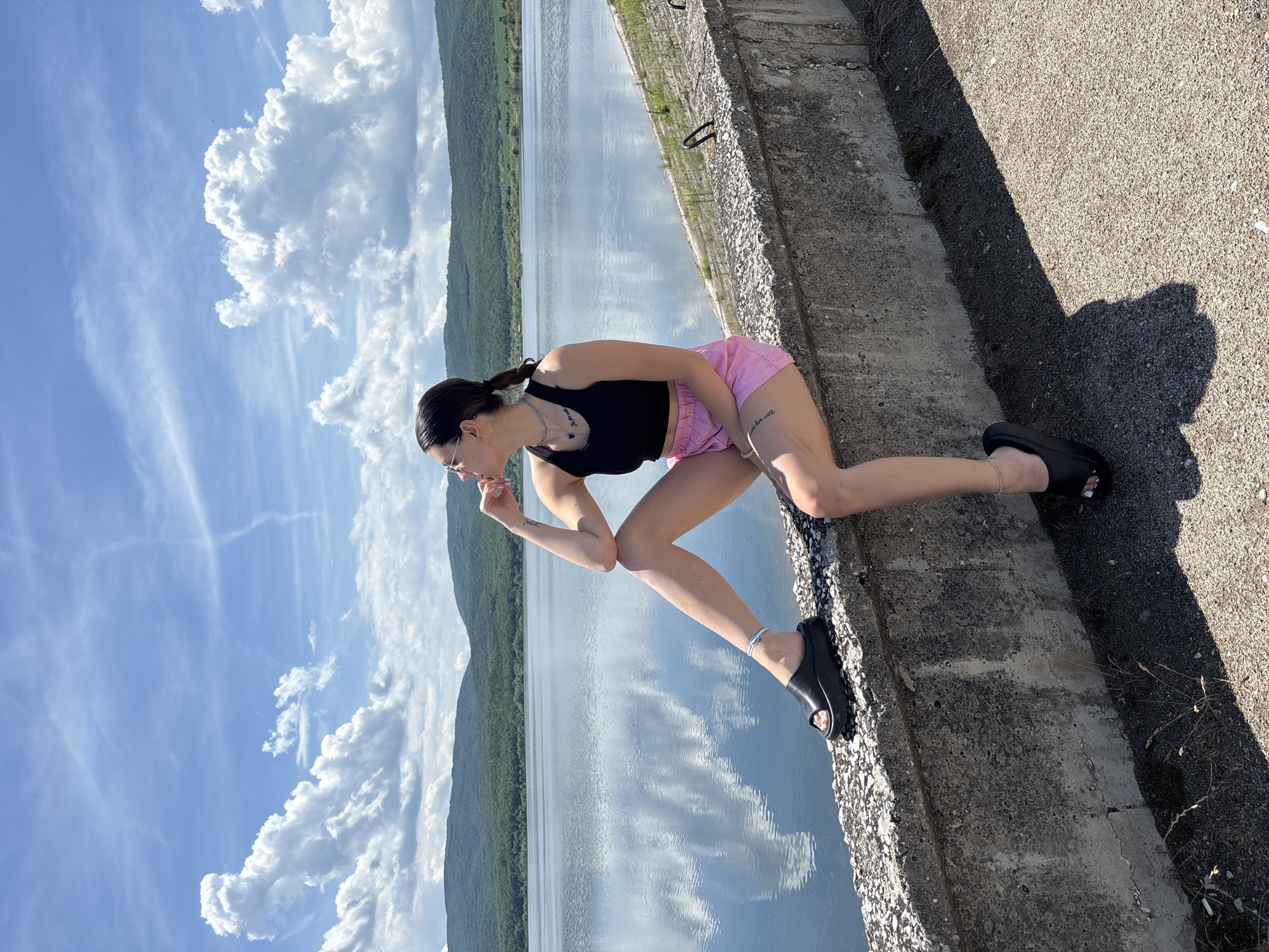 A young woman is captured in a posed shot, sitting on a concrete ledge overlooking a vast body of water. Her dark hair is pulled back in a ponytail, and she wears glasses. She is dressed in a black sleeveless crop top and pink checkered shorts. Her left arm is bent, with her hand near her chin, while her right arm rests on her thigh. She has a silver-colored anklet on her right ankle and a thin necklace with small blue charms. A tattoo on her right thigh reads "live her will". She is wearing black, open-toed slide sandals.
The setting is a scenic outdoor location, specifically in Ognyanovo, Bulgaria. Behind the woman, a wide expanse of calm water reflects the bright blue sky and fluffy white clouds. In the distance, lush green mountains or hills rise along the horizon, framing the water. The sky is largely clear with scattered cumulus and cirrus clouds, indicating fair weather. The bright sunlight suggests it is daytime, possibly late morning or afternoon, with strong shadows visible on the concrete surface to the right. The woman's pose appears relaxed and contemplative as she looks slightly downwards or to the side.