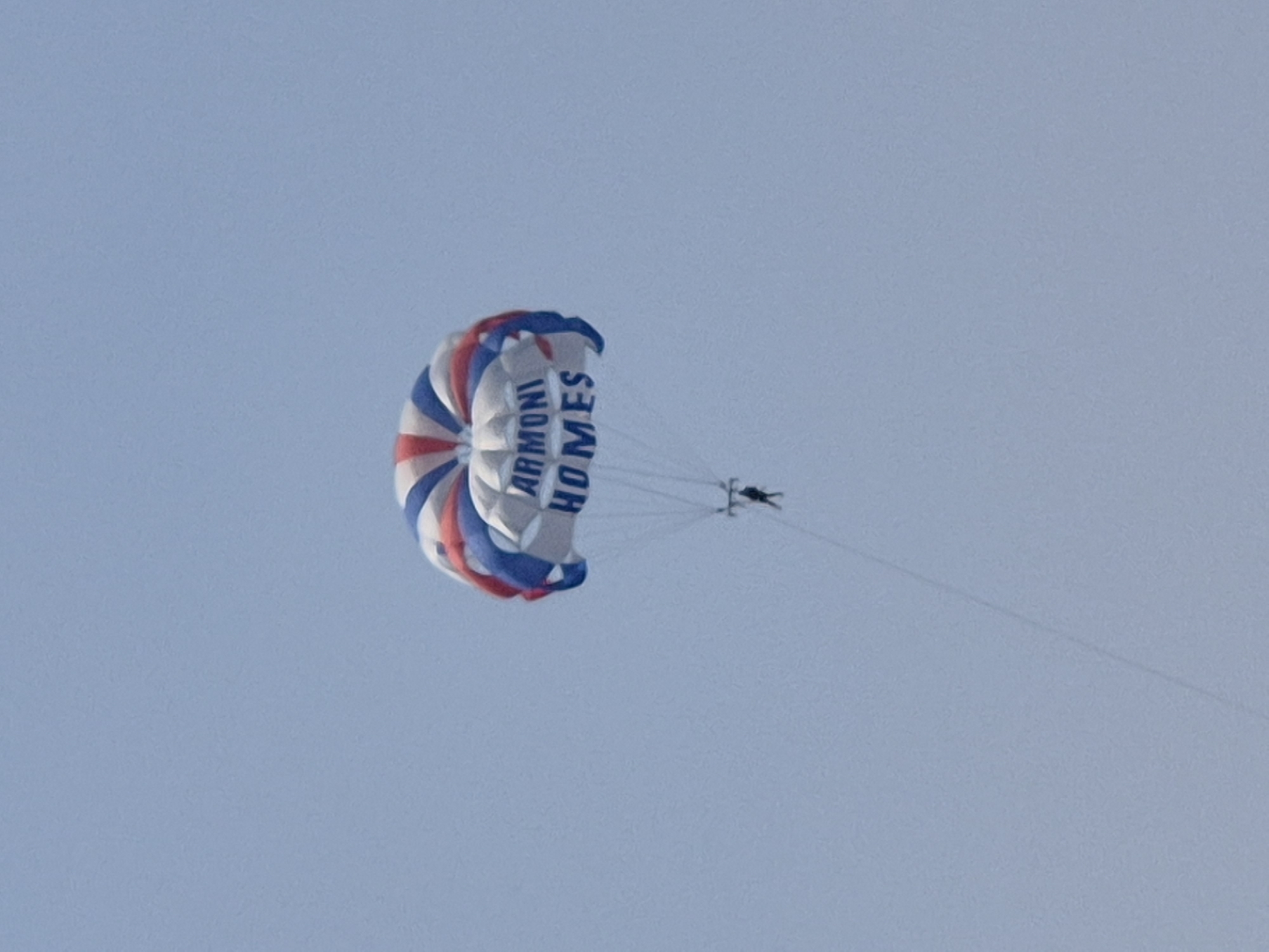 A person is parasailing with a red, white, and blue parachute against a clear sky. The parachute has "ARMONI HOMES" written on it in blue lettering. The person is suspended from the parachute by lines. The image appears to be taken from a distance, looking up at the parasailer. The sky is a light blue and shows no clouds.