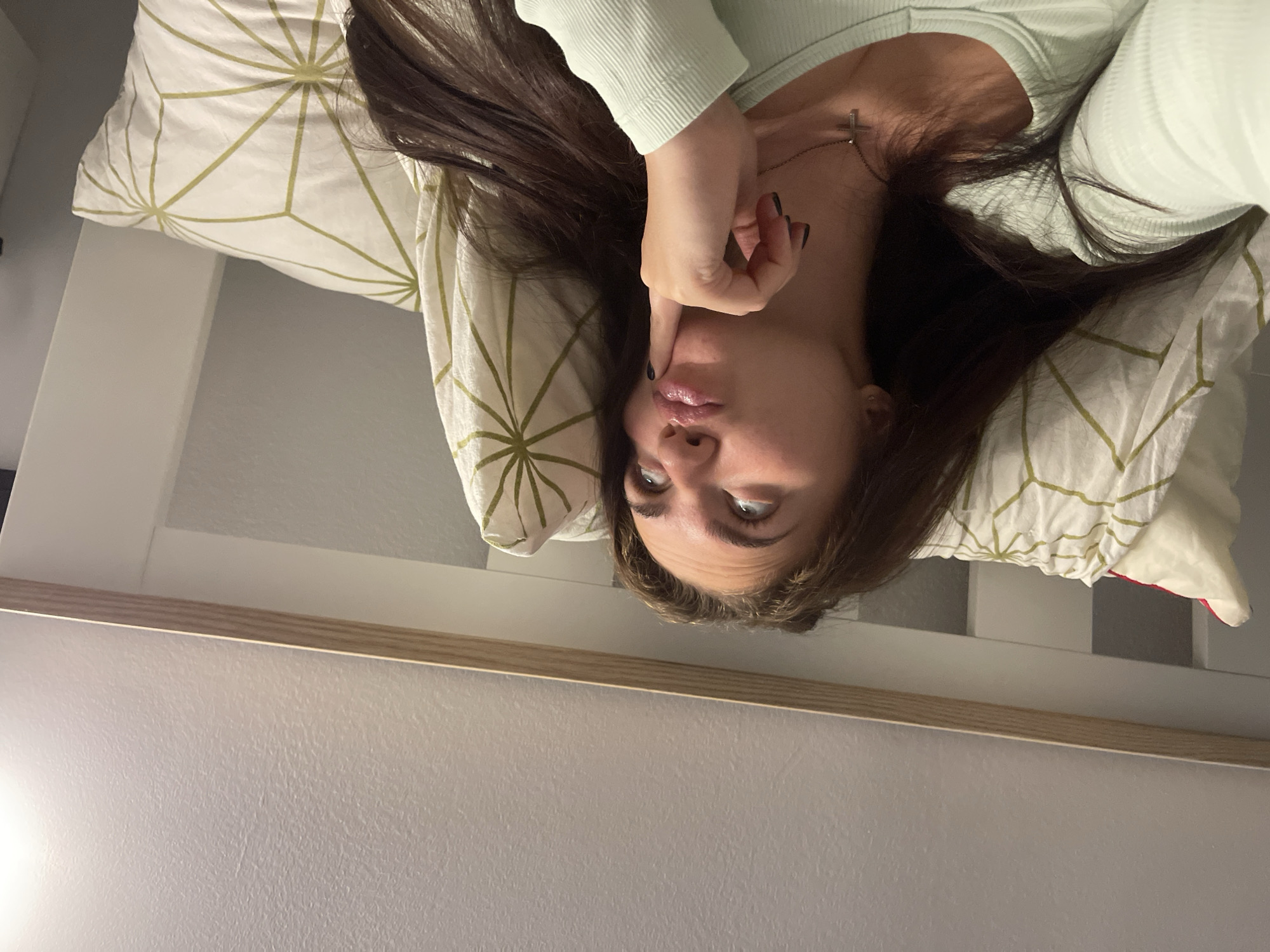 The image shows a young woman lying in bed, looking upwards with her finger on her chin in a pensive pose. She has long, dark hair and is wearing a light green ribbed top and a silver cross necklace. The bed has white pillows with a geometric pattern of green lines on a white background. The headboard of the bed is white with a wooden trim. The ceiling is textured and appears to be painted white. The lighting suggests an indoor setting, likely a bedroom, during the evening or night. There is no information in the image to indicate the location in the city of Мосметаллоконструкция Онт, Russia.