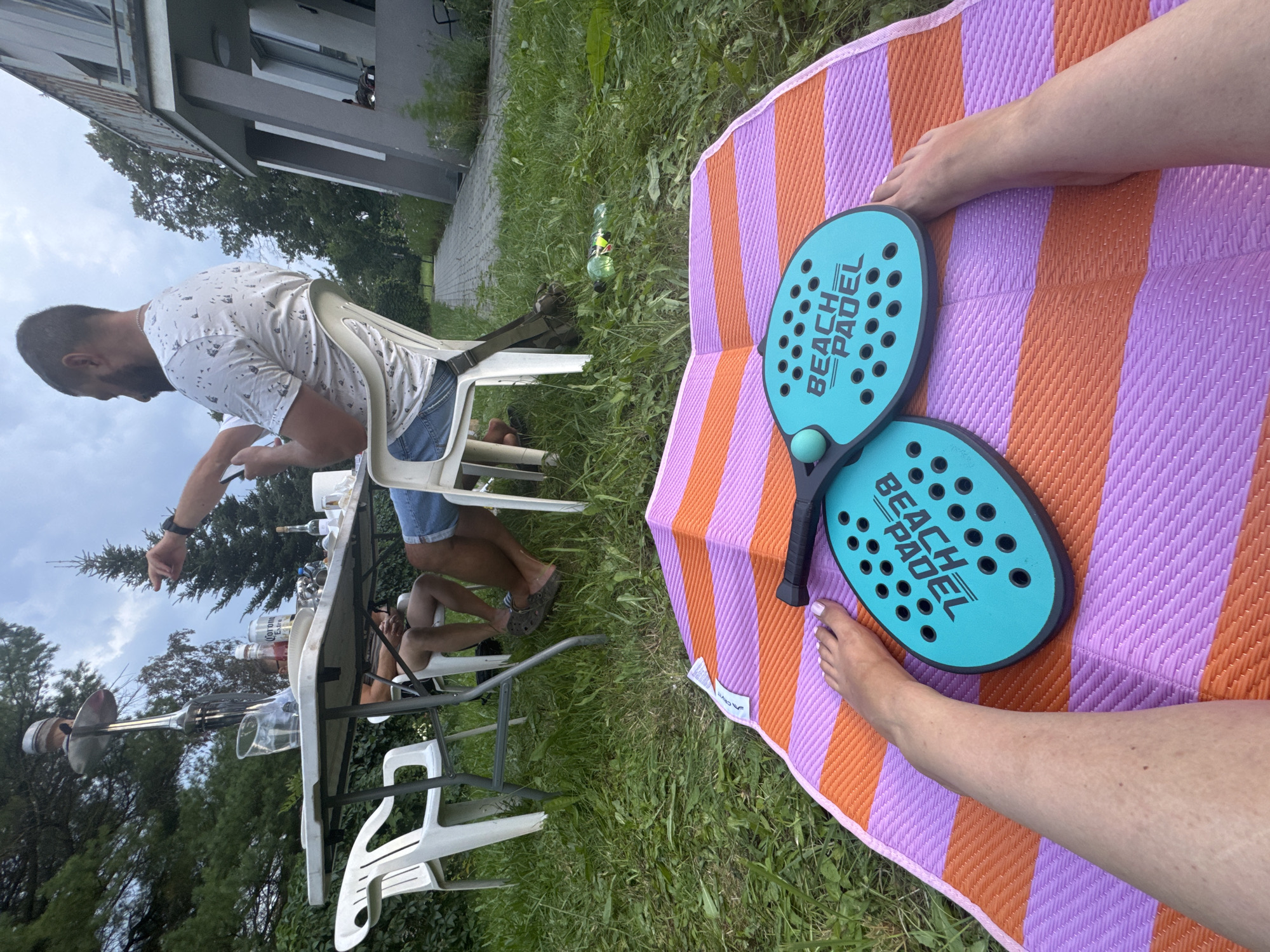 In an outdoor setting, likely a backyard, two teal colored beach paddleball rackets are laid out on an orange and purple striped mat. One racket has the text "BEACH PADEL" printed on it. A person's bare feet are visible on the mat, with one foot positioned near the rackets. In the background, a group of people are seated around a table, with drinks and food present. A hookah pipe is visible on the table. The environment includes green grass, trees, and a building in the distance, suggesting a residential area in the city of Katowice, Poland. The sky is overcast.