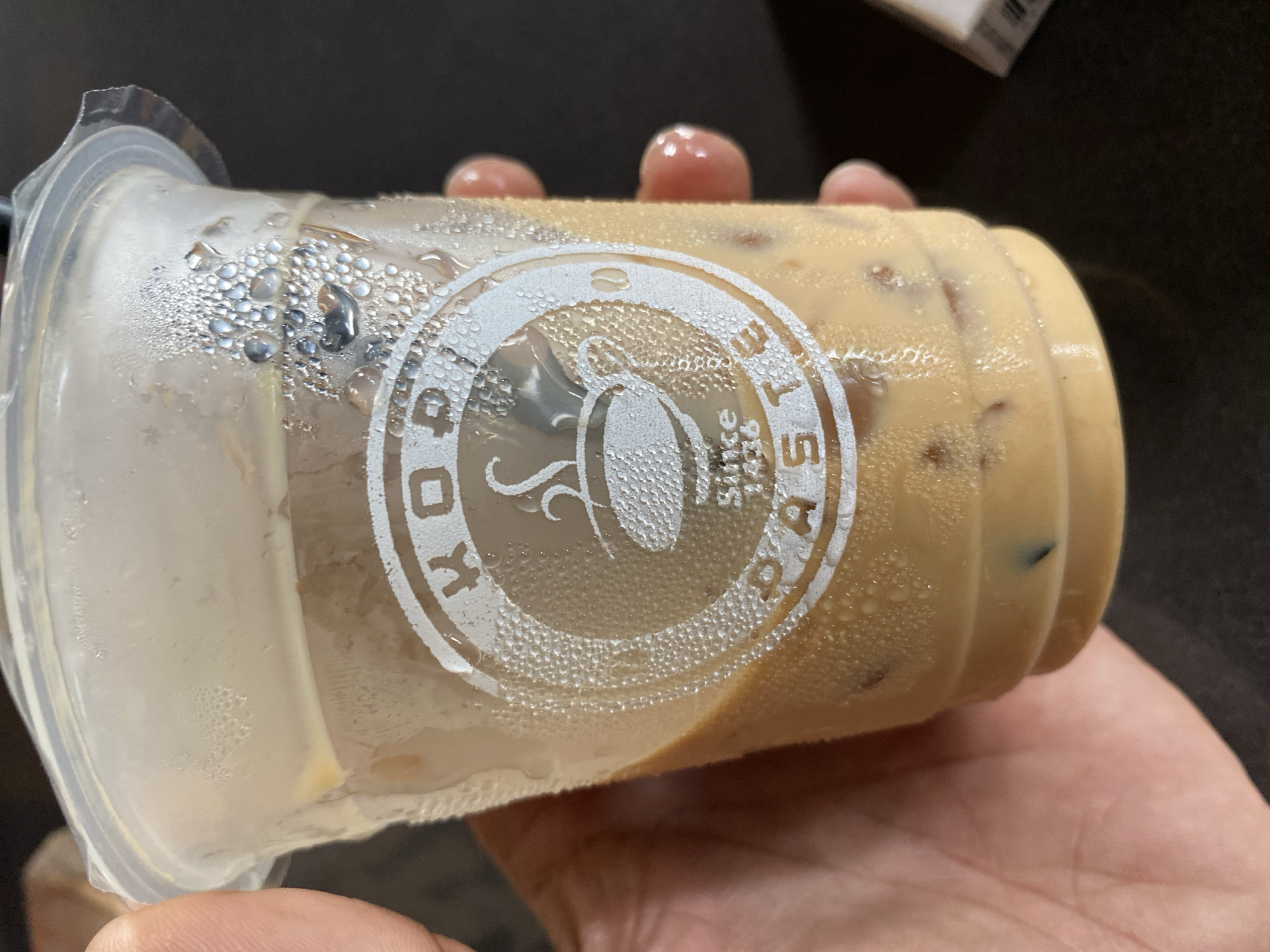 A close-up, horizontal shot shows a person's hand holding a clear plastic cup filled with an iced, light brown beverage, likely a milky coffee. The cup is wet with condensation, indicating a cold drink in a warm or humid environment. Several fingers are visible gripping the cup, with a thumb supporting it from below.
The cup features a white, circular logo prominently displayed on its side. The logo reads "KOPI" on the left and "PASTE" on the right, encircling a stylized image of a coffee cup with steam rising from it. Below the coffee cup icon, smaller text reads "since 1986". Ice cubes are visible within the beverage, which has a smooth, creamy appearance. The background is dark and out of focus, providing no specific details about the immediate surroundings beyond suggesting an indoor setting. The image captures the act of holding a refreshing drink, possibly consumed in Tanjongkapai, Indonesia.