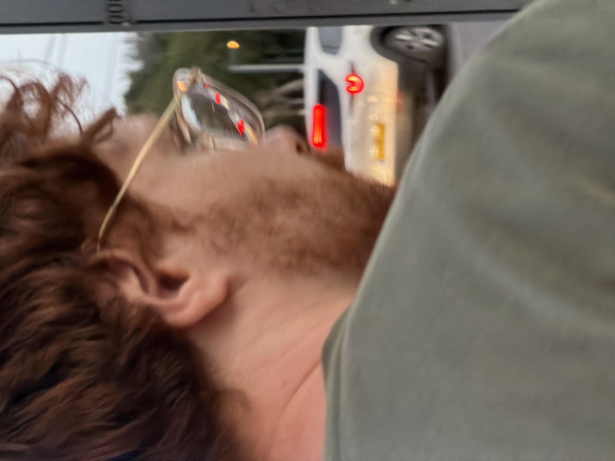 A close-up, rotated image shows a man with reddish-brown hair and a beard, wearing glasses. He is positioned near a window of what appears to be a vehicle, judging by the blurred exterior view through his glasses reflection. The reflected image shows what seems to be a car and road signs, suggesting an urban environment. Given the instructions, it is hypothesized that this image was taken somewhere in Tel-Aviv, Israel. The man is wearing a light green shirt, and he seems to be looking upwards, out the window.
