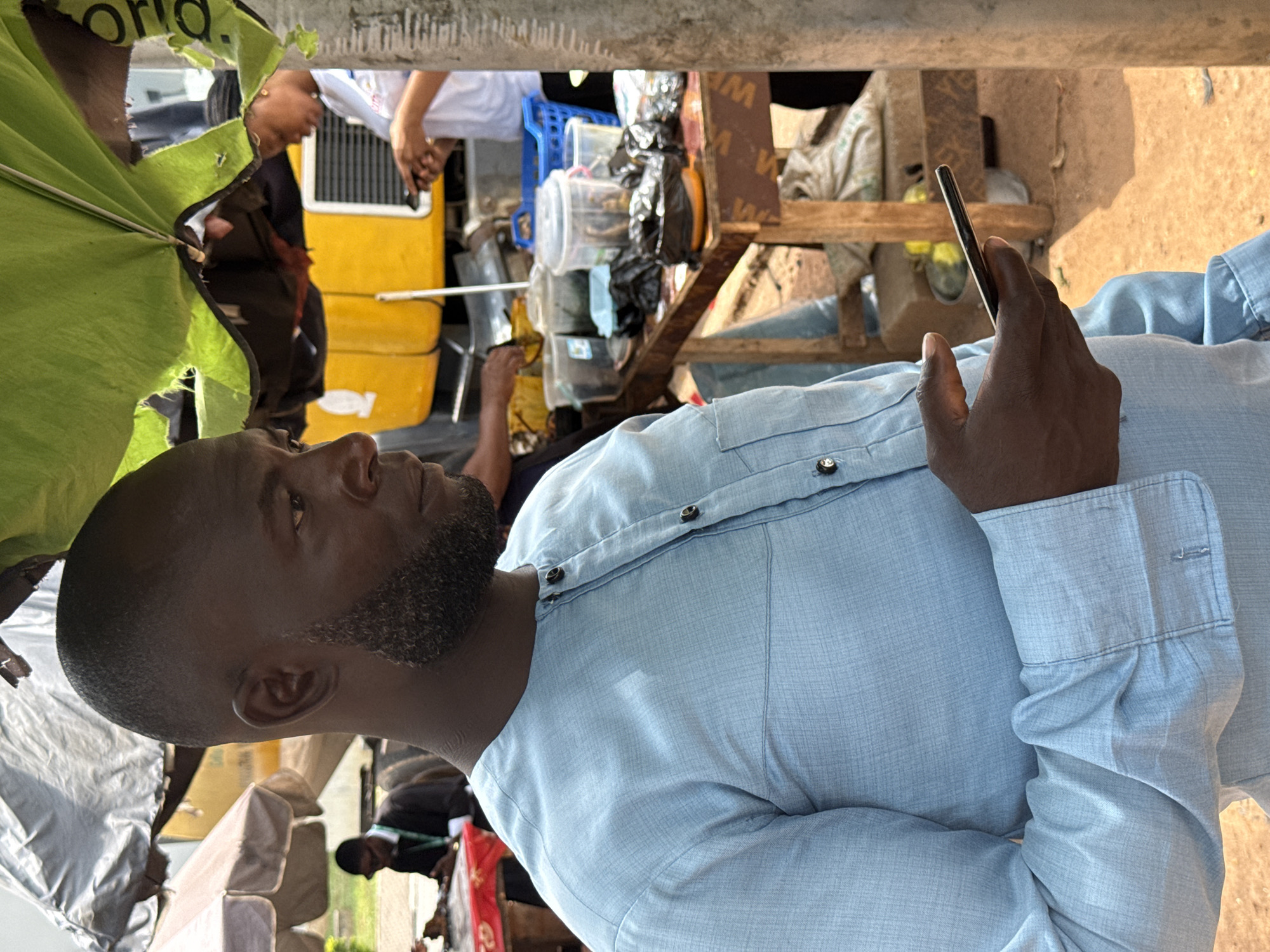 The image depicts an outdoor scene, likely a market stall, in Abuja, Nigeria. A man of African descent, wearing a light blue traditional Nigerian garment with dark buttons, is the primary subject. He has a close-cropped haircut and a well-trimmed beard. He is holding a smartphone in his right hand, looking towards the side with a neutral expression.
In the background, a bright yellow vehicle, possibly a bus or a truck, is visible. A woman wearing a white top is standing near the vehicle, also looking at a phone. Several tables are set up with various items, including plastic containers filled with food or goods, plastic bags, and a blue plastic basket. Some of these items appear to be for sale.
Overhead, part of a green and yellow umbrella is visible, providing shade. The ground appears to be a dusty, reddish-brown color, typical of dry earth. Other people are partially visible in the background, suggesting a bustling environment. The overall impression is that of a lively street scene in Abuja.