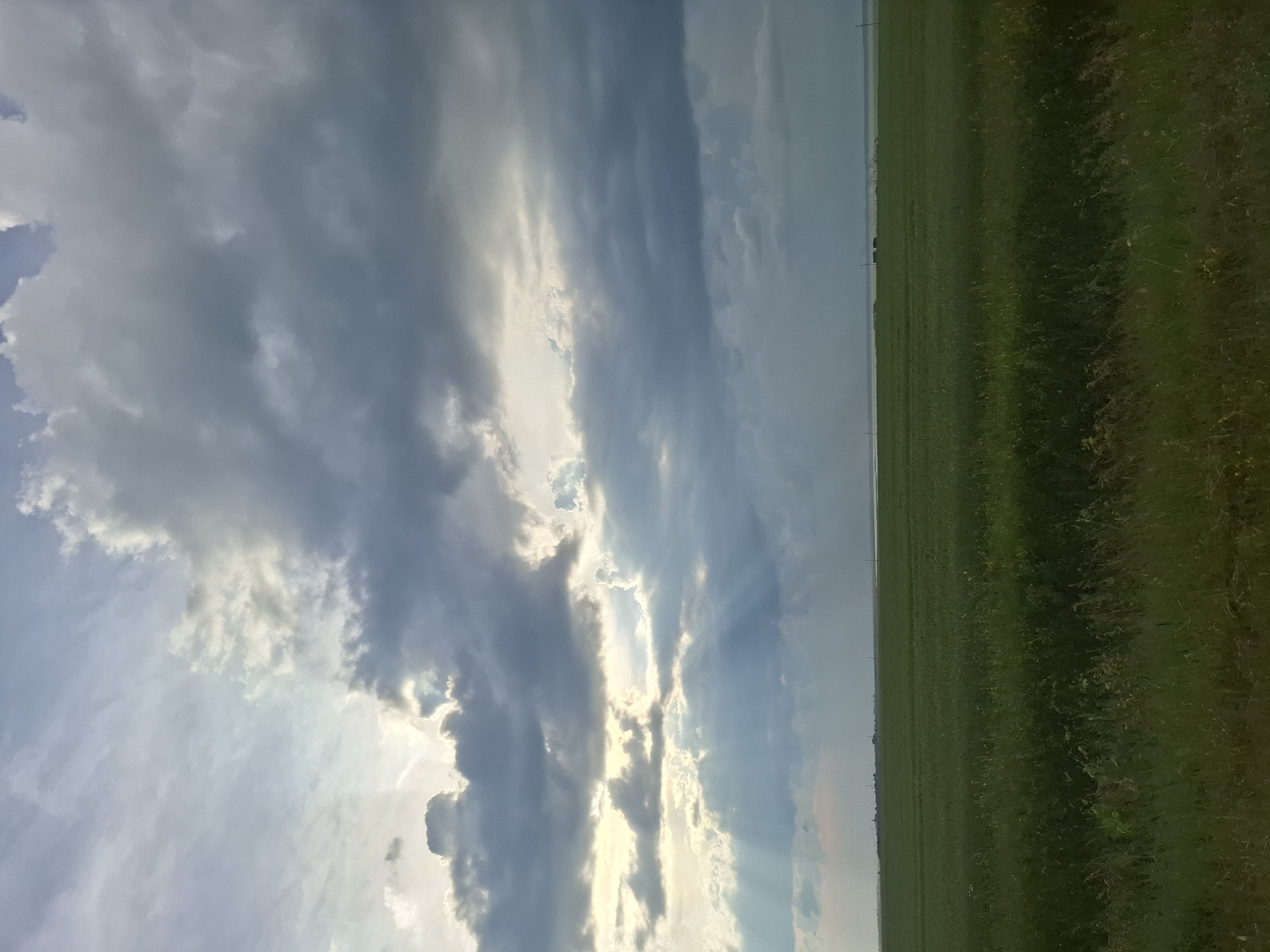 This is a landscape photograph taken in a rural area. The image is dominated by a dramatic sky filled with a mix of dark, brooding clouds and patches of bright sunlight that create visible sunbeams. The clouds suggest that the weather may be changing, possibly with the approach of a storm or the clearing after one.
To the right of the sky, a vast, flat expanse of green grassland stretches towards the horizon. The grass appears to be a field, possibly used for agriculture or as pasture. The horizon line is sharp and well-defined, separating the sky from the land. In the far distance, faint vertical lines suggest the presence of utility poles or fence posts, indicating human presence in the otherwise natural landscape.
The orientation of the photograph is such that the sky takes up the majority of the left side and the grassland occupies the right side. The lighting suggests it is either early morning or late afternoon, given the angle and intensity of the sunbeams. There are no people or man-made objects prominently featured, emphasizing the natural scenery. The overall mood of the image is one of natural grandeur and the vastness of the open landscape. The location context indicates this scene is from the Avangard rural district in Kazakhstan.