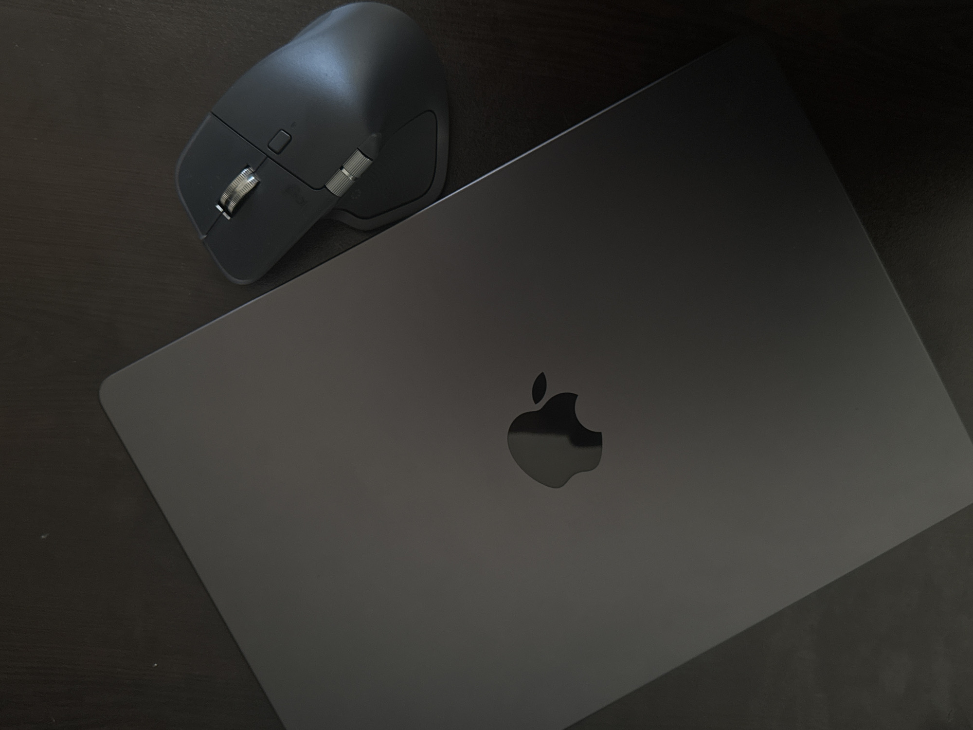 A gray laptop with an Apple logo on the closed lid is on a dark surface. A gray wireless mouse sits to the left of the laptop. The environment appears to be a dark, possibly wooden desk or table. There is no evidence of the location being Sofia, Bulgaria.