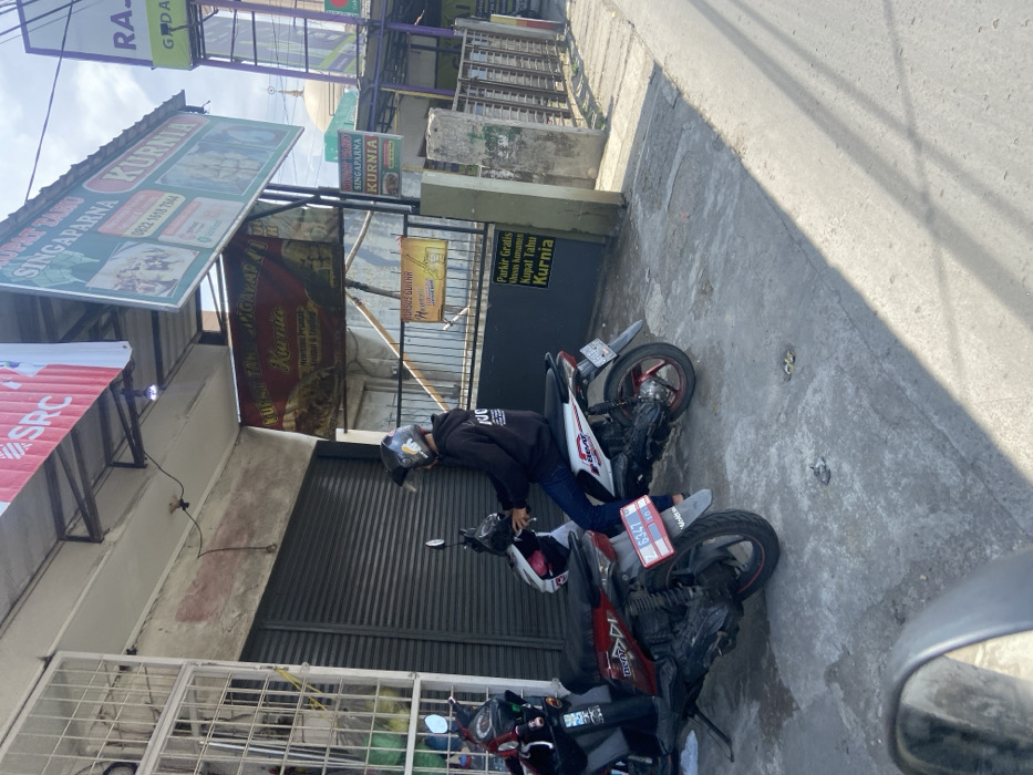 The scene takes place in front of a commercial building on a street in the city of Kecamatan Dayeuhkolot, Indonesia, located at latitude: -6.965, longitude: 107.593. A young male is sitting on a white motorcycle, wearing a black hoodie with a red and white logo, blue jeans, and a white helmet. The motorcycle has a red license plate that reads "6327," which is a local Indonesian license plate. The motorcycle is parked on the sidewalk in front of a building with an awning that has a sign written in Indonesian. The building also has several other signs, some of which have prices and information about the store. The signs are all in Indonesian. The street is paved and grey, with white lines. The building has a closed metal gate. The sidewalk is narrow, and there is some vegetation growing between the sidewalk and the street. The sun is shining, and there is a shadow on the street. The sky is clear and blue. The store sign appears to advertise a Singaparna Kurnia restaurant, which likely serves local Indonesian cuisine.