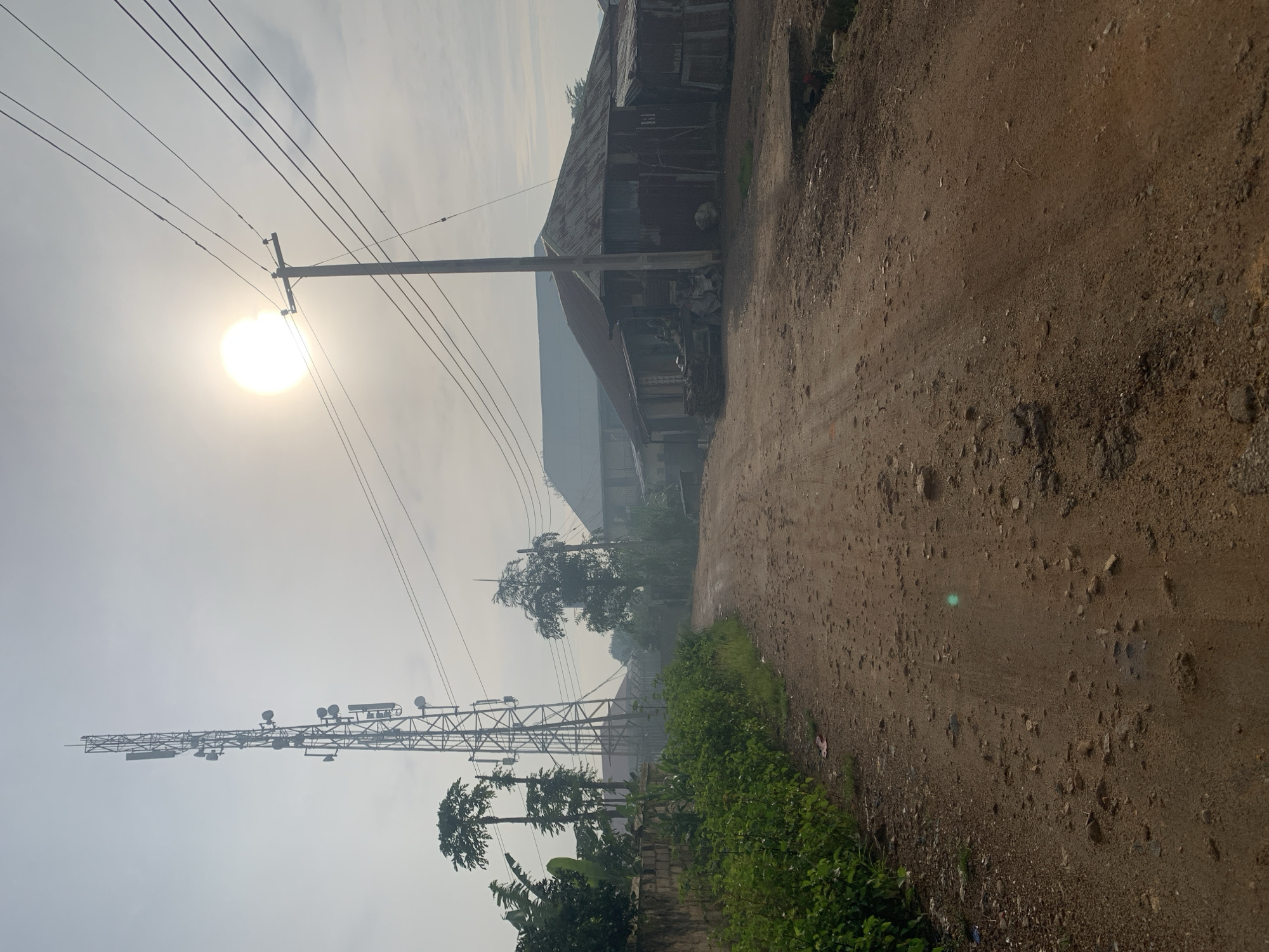 The image depicts a rural or semi-urban scene, likely in Rafin Par, Nigeria, on an overcast day with the sun partially obscured. A dirt road, rutted and uneven, leads into the distance. On the left side of the road, lush green vegetation grows along a low stone wall, with several tall trees, including what appear to be papaya trees, rising above it. Dominating the left side of the frame is a tall, lattice-style telecommunications tower equipped with multiple antennas and dishes.
On the right side of the road, several structures are visible. The foreground features a long, low building with a corrugated metal roof, appearing weathered and somewhat dilapidated. Further down the road, a larger building with a similar roof is partially visible, alongside more vegetation and a utility pole supporting electrical wires. The sky is hazy, with the sun casting a diffused glow. There are no people or animals clearly visible in the image. The overall impression is one of a developing area with basic infrastructure.