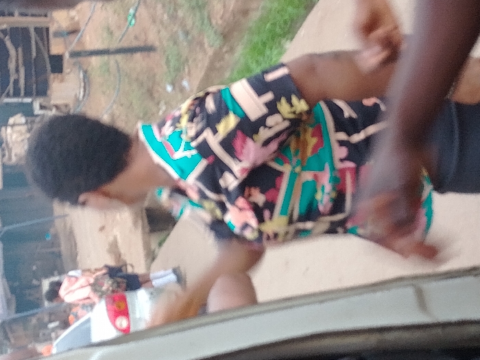 The image portrays an outdoor street scene in Orie Uratta, Nigeria. In the midground, a person with short, dark hair, seen from behind and facing slightly right, wears a short-sleeved shirt patterned with black, teal, pink, yellow, and white geometric and abstract shapes, over dark trousers. Their right arm is partially extended forward. In the lower right foreground, two dark-skinned hands are visible, with one appearing to interact with the main subject's right arm or hand area.
To the left, the side of a white vehicle, possibly a car, is partially visible, displaying a red taillight or reflector. Further in the background, another individual is seen walking away, wearing a light-colored top (possibly peach), dark shorts, and white socks, carrying a patterned backpack. The immediate ground consists of a light-colored, paved surface, likely a sidewalk. To the right of this path, a concrete drainage channel is visible, bordered by green vegetation. The background features blurred structures consistent with an urban or semi-urban environment, including what appears to be corrugated metal and dark poles. The scene is illuminated by ambient daylight.