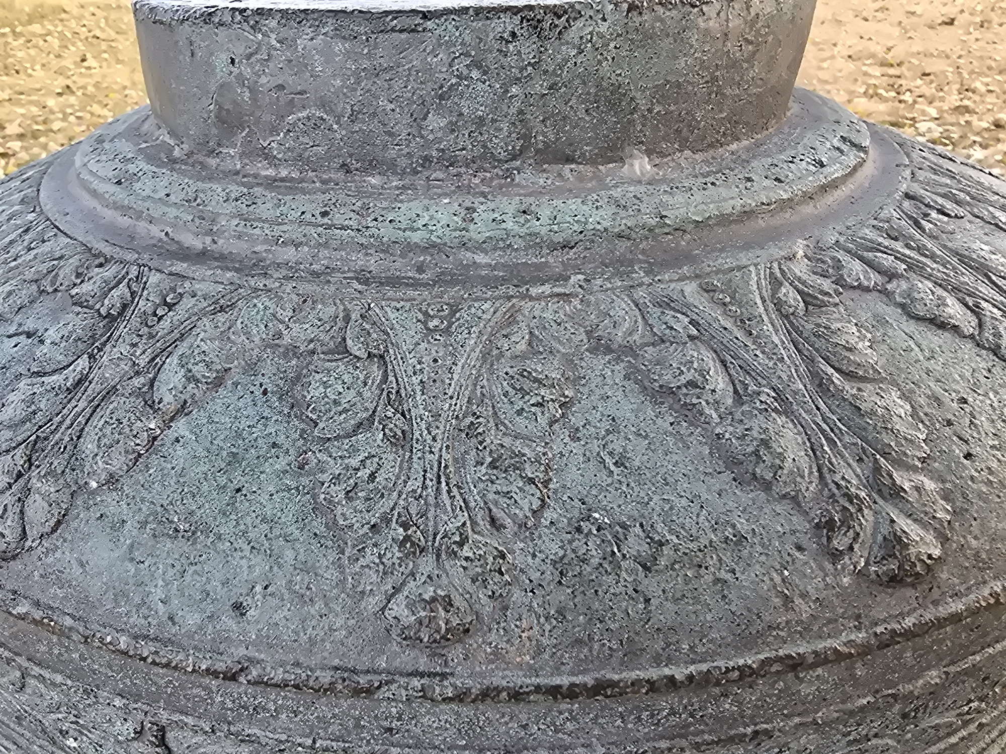 The image shows a close-up, detailed view of the weathered, patinated surface of a large, possibly bronze, object. The object has a rounded, bell-like shape with a distinct rim or collar near the top. The surface is adorned with intricate, raised relief carvings that appear to be floral or foliate motifs, with detailed leaf and stem patterns. The material exhibits a greenish patina, indicative of age and exposure to the elements, possibly at a historical site. The background is out of focus and appears to be a granular, sandy or gravelly surface, suggesting an outdoor environment. This object is located at the Rubenshuis museum in Antwerp, Belgium, specifically it is part of the "Carillon" sculpture by Lucassen, situated in the museum's garden.