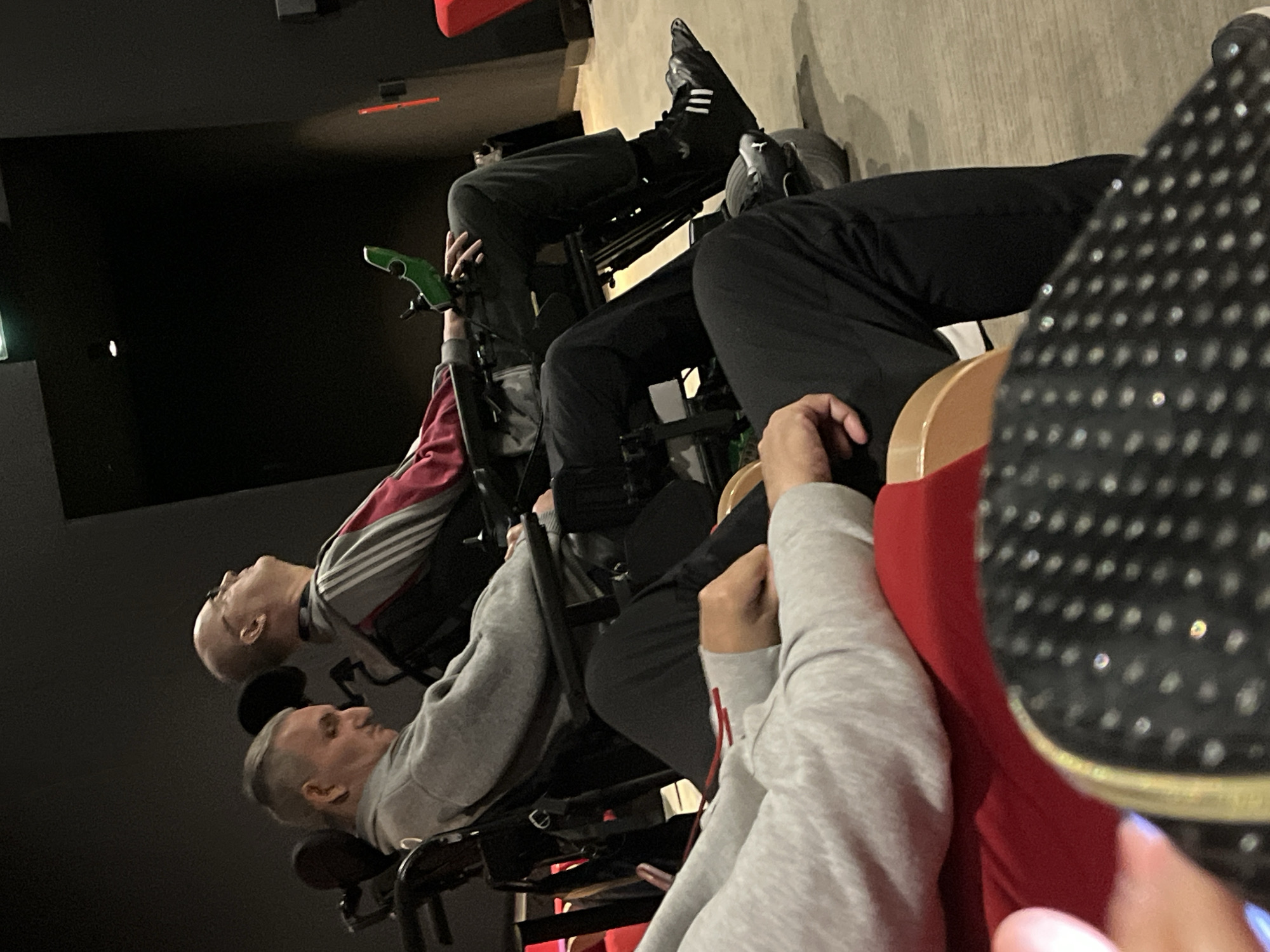 In an indoor setting resembling a theater or auditorium, two individuals are seated in wheelchairs. The person closest to the camera is wearing a grey sweatshirt, while the other is in a grey and red jacket. Both appear to be looking toward a dark screen. A third person, partially visible, is wearing a red shirt with a grey sleeve. The foreground includes a black shoe with silver embellishments. The location appears to be Montbrison, France.