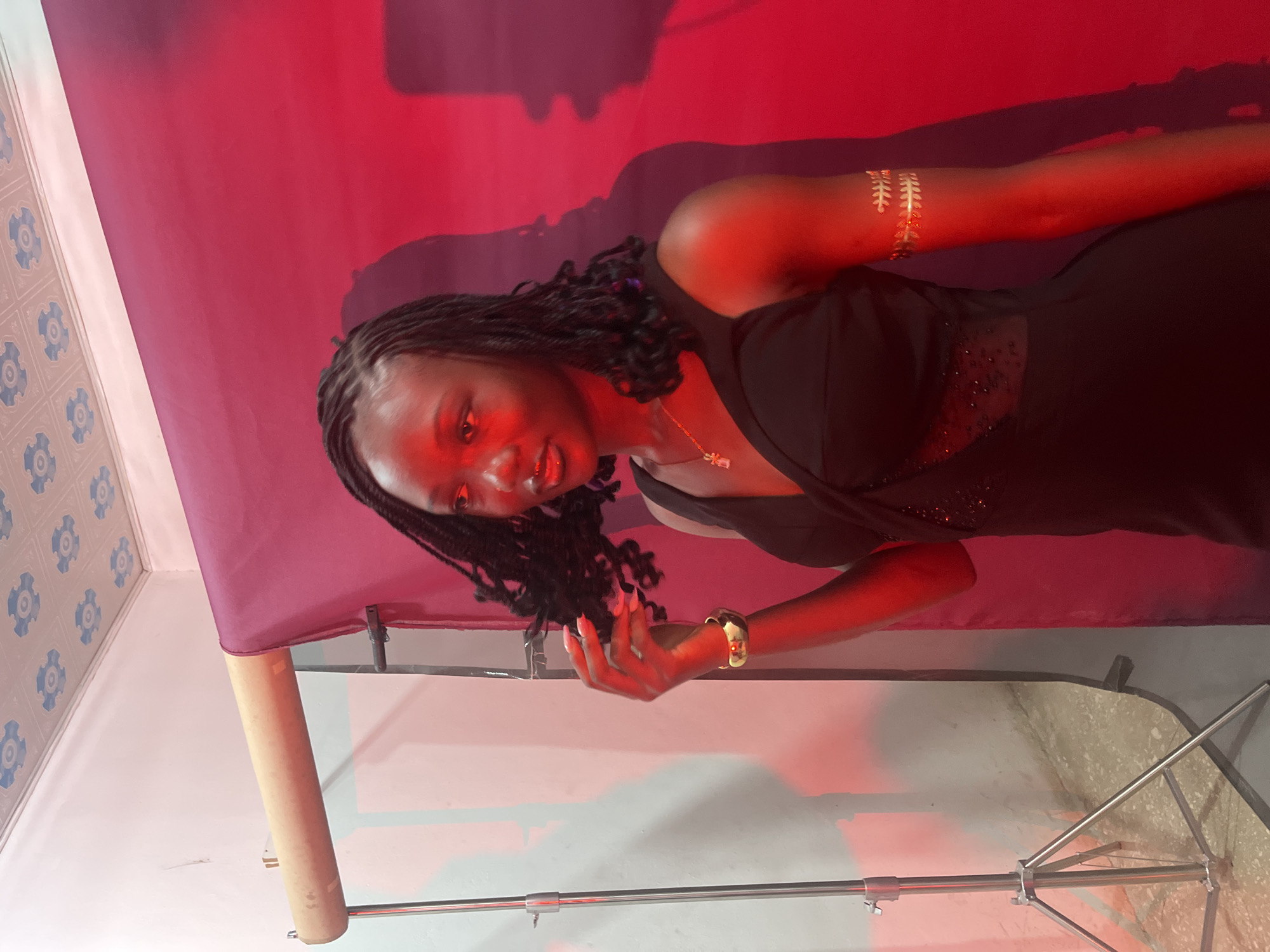A young Black woman with braided hair is posing for a photo against a red backdrop in what appears to be a studio or prepared photo area. She is wearing a sleeveless black dress with sheer, bejeweled panels on the side. She is accessorized with a gold bracelet on her left wrist and a gold arm cuff on her right upper arm. Her left hand is near her face, touching her hair. The lighting is predominantly red, casting a warm glow and creating shadows on the backdrop and wall. A lighting stand is visible to the left of the woman, and a roll of paper or fabric is mounted on a stand behind her. The ceiling has a decorative pattern. Based on the context provided, the location is Gura Top, Nigeria.