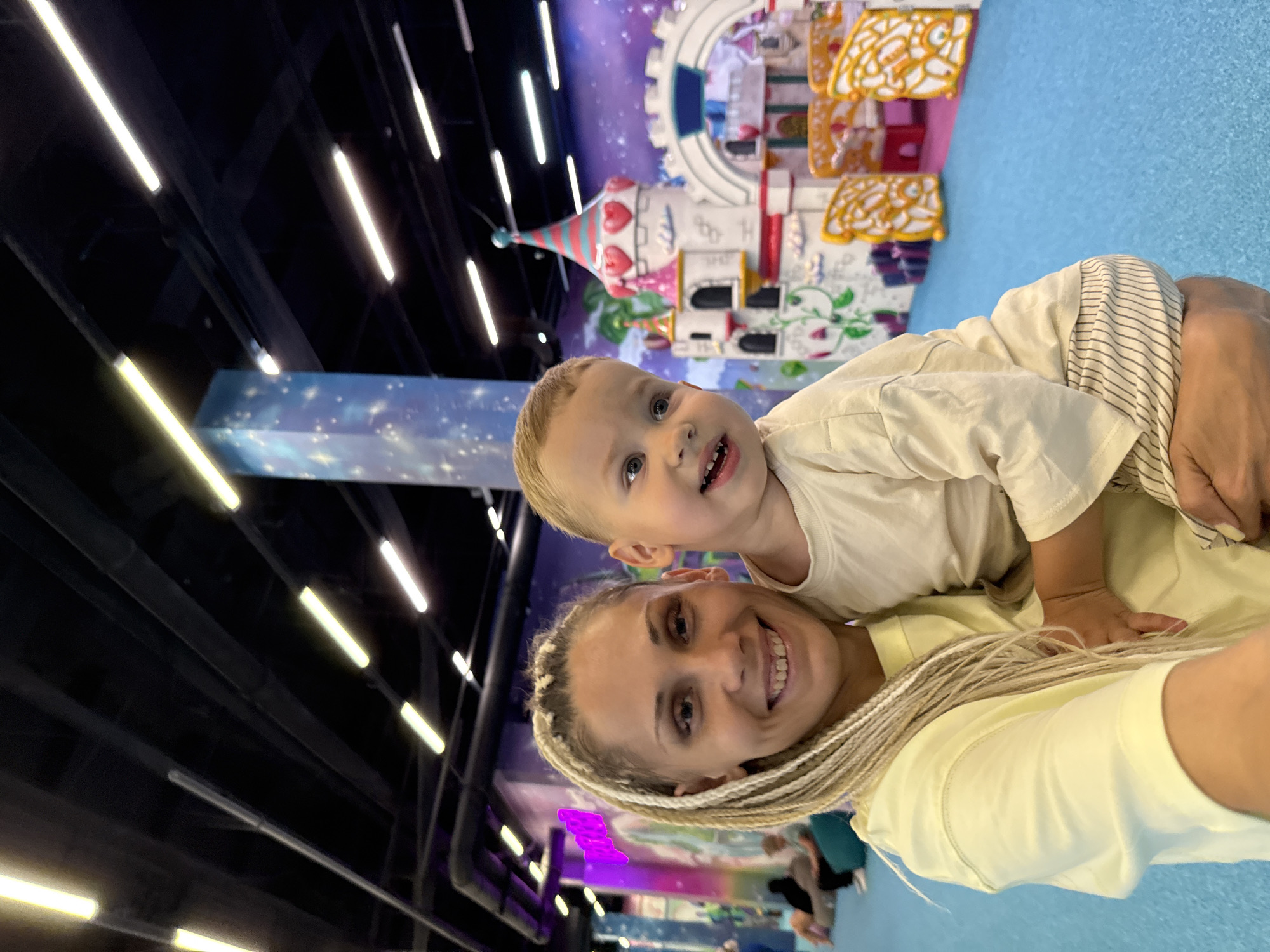 A blonde woman with braided hair smiles as she holds a young boy in front of a colorful indoor playground. The playground features a large, pink and white castle with heart-shaped windows and flags, as well as yellow and orange play structures. The background is painted with a purple, starry sky motif. The woman is wearing a yellow t-shirt, and the boy is in a cream-colored shirt and striped pants. The floor is bright blue. The scene appears to be from a children's entertainment center or amusement park. The image does not provide any information to suggest the location is in the city of Zhukovo, Russia.