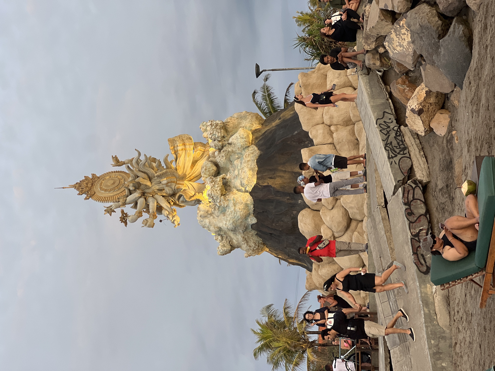 The scene depicts a public gathering area at dusk or early evening in Kerobokan Kelod, Indonesia, under an overcast sky. A large, multi-armed golden statue, possibly a deity, serves as the central focal point, illuminated by spotlights. It stands atop a dark, conical base surrounded by lighter, irregular rock-like formations.
In the foreground, on sandy ground, a person wearing a baseball cap, sunglasses, and a dark top reclines on a green lounge chair, actively looking at a mobile phone. A green coconut drink rests on a small, dark surface next to the chair.
Immediately behind this, a series of broken concrete slabs, marked with graffiti, form part of the terrain leading to the statue. Large, grey and brown rocks are scattered across the sandy ground.
Numerous individuals are present throughout the mid-ground. From left to right:
* A man in a black shirt and khaki shorts walks with a woman in a black dress, both appearing to hold hands.
* Near the base of the statue, several people are standing and conversing. One man in a red shirt and a grey fanny pack is eating a cob of corn.
* A woman in a black swimsuit and white headband stands alone, observing the surroundings.
* Further to the right, multiple individuals are seated on the large rocks, some looking at mobile phones.
In the background, on both the left and right sides, green palm trees are visible. A black street light pole stands on the right. The overall environment suggests a waterfront or beach-adjacent public space with recreational activity.