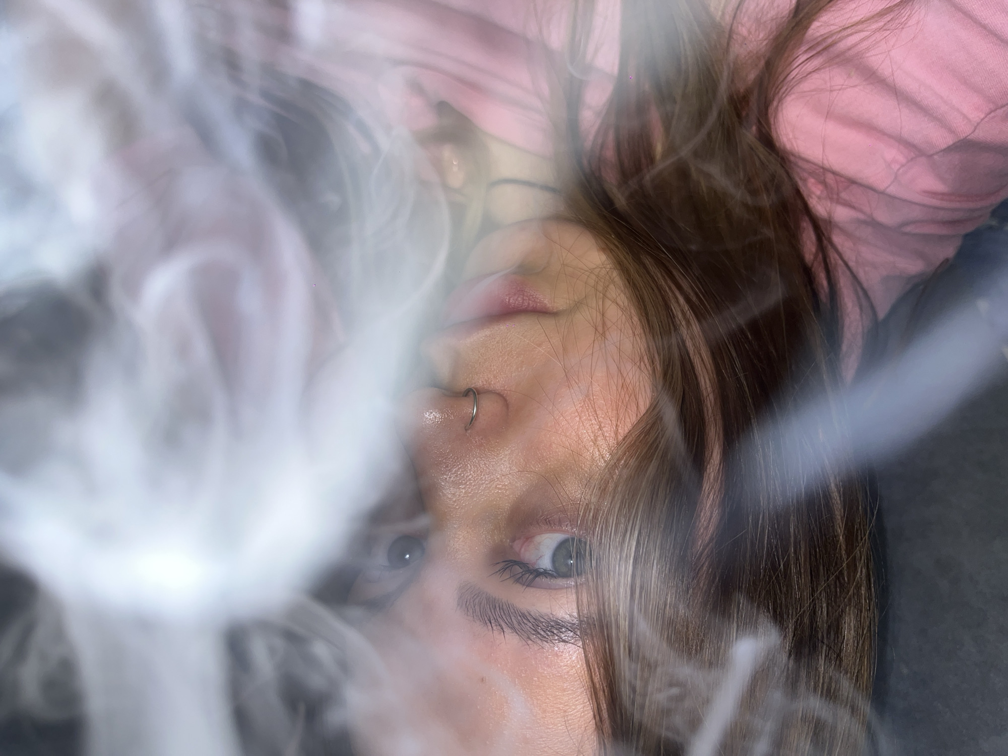 The image shows a close-up, head-and-shoulders shot of a young person lying down, with their head tilted to the side and their gaze directed upwards. A swirling mist or smoke obscures parts of the face and hair, creating a dreamlike or ethereal atmosphere. The person has dark brown hair, visible eyelashes, an eyebrow piercing, and appears to be wearing a pink garment. The background is dark and out of focus. There is no indication of the location being in Novopokrovskaya, Russia; the image provides no geographical context.