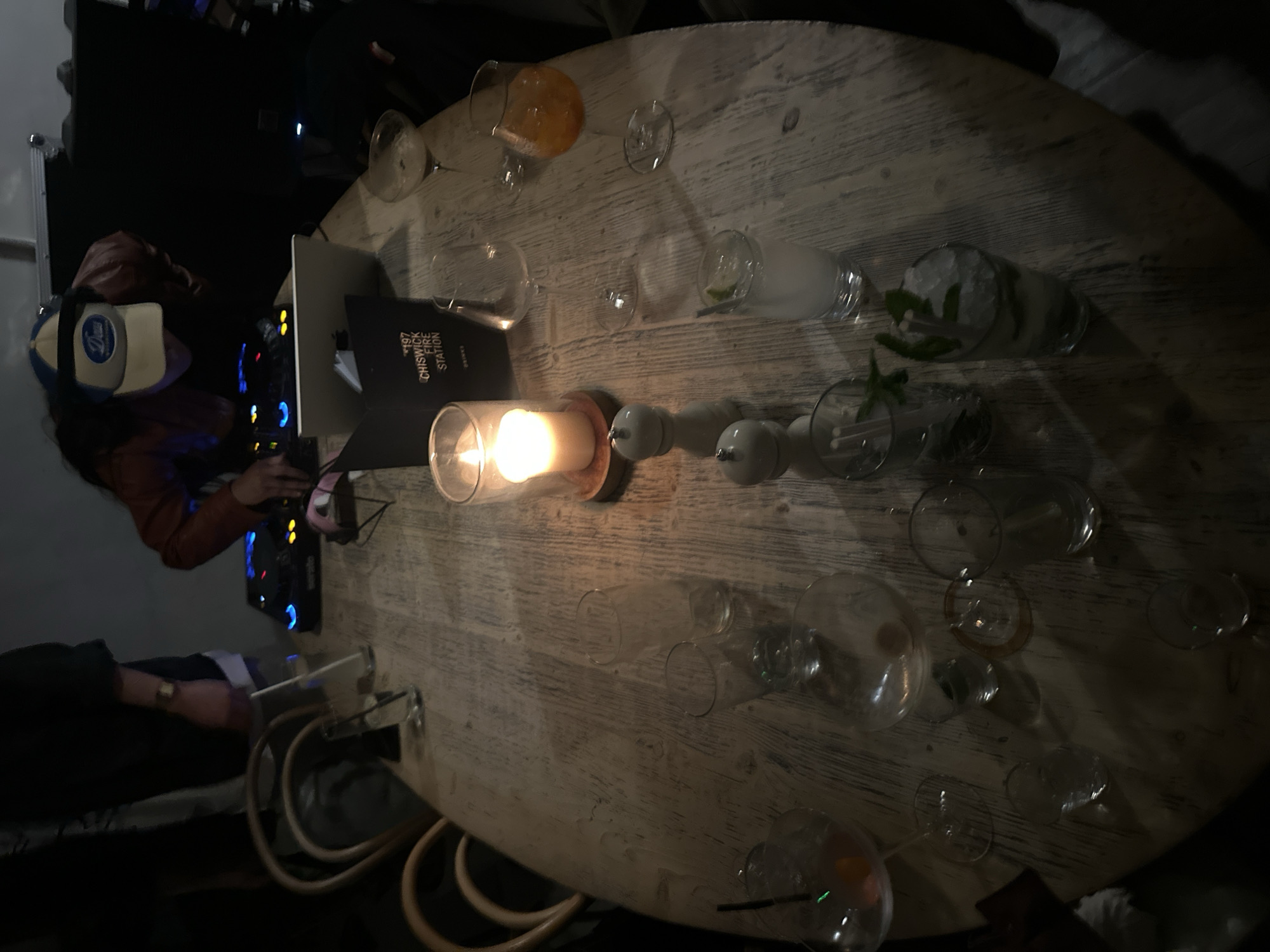 The image shows an oval wooden table set with numerous empty glasses, cocktail glasses containing beverages, a candle in a glass holder, and salt and pepper shakers. A DJ wearing a baseball cap and a brown jacket is operating DJ equipment including a laptop and mixing console. A menu with the words "197 Chiswick Fire Station" is visible near the candle. Another person's arm is visible to the bottom left of the table, wearing a watch and dark clothing. The location is likely Chiswick Fire Station, London, United Kingdom.