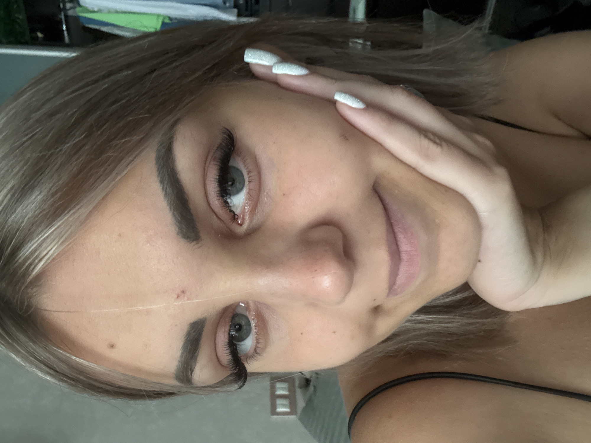 A close-up, selfie-style photograph shows a young woman with blonde hair and light-colored eyes. She is looking at the camera and resting her right cheek on her left hand, which has white, sparkly nails. Her left eyebrow appears to have a small blemish. The background is blurred but appears to be an indoor setting, possibly a bedroom with a closet containing clothing. There is no indication of the city of Yuzhno-Sakhalinsk, Russia.