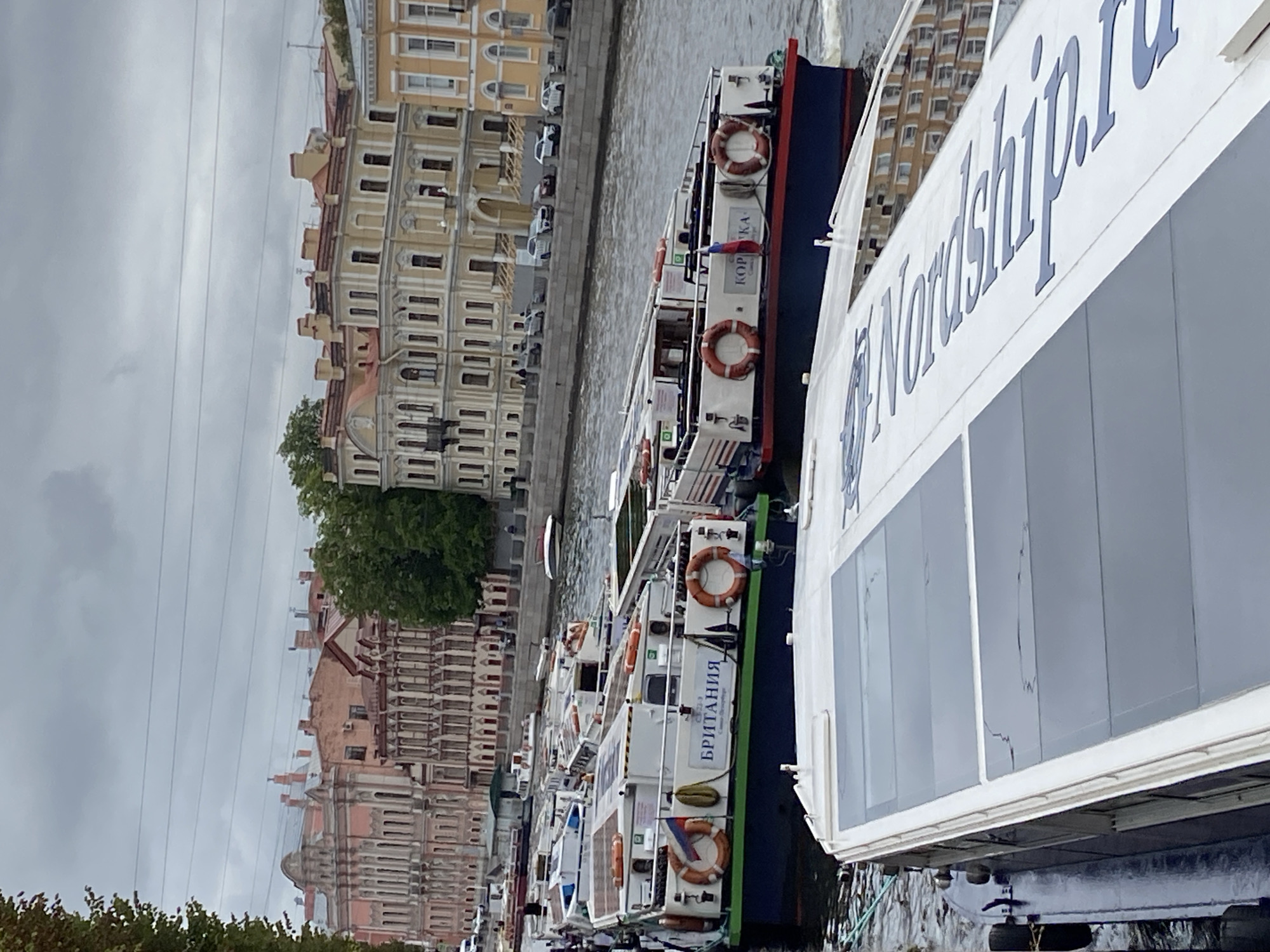 The image depicts a cloudy day in Saint Petersburg, Russia, with multiple tour boats docked along a canal or river. In the foreground, a white tour boat with a dark blue hull is partially visible, its roof featuring tinted glass panels. Behind it, a line of several similar tour boats are moored together. One of these boats prominently displays the Cyrillic text "БРИТАНИЯ" (Britannia) and a Russian flag. Another boat to its right shows the Cyrillic text "КОРКА" (Korka). The background reveals a street with cars and a row of classical European-style buildings, characterized by ornate facades, multi-story structures, and pitched roofs, some with visible chimneys. Trees line the canal bank. The overall scene suggests a typical tourist activity in the historic city center of Saint Petersburg.