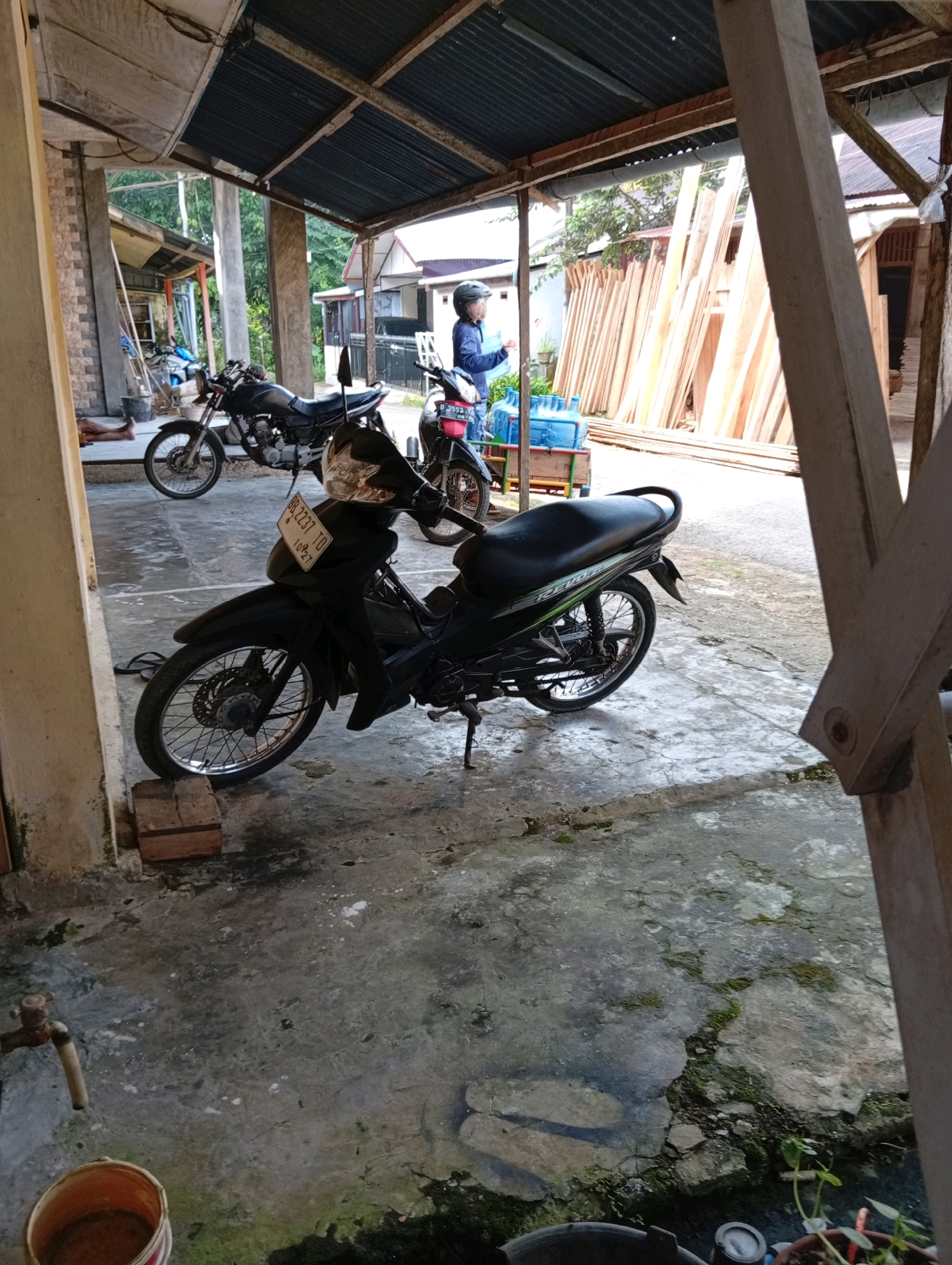 A motorcycle is parked under a rustic shelter with a corrugated metal roof. The motorcycle is black with green accents and a license plate that reads "BB 2237 TD". A person wearing a blue shirt and a helmet is standing next to the motorcycle, looking to the left. In the background, there are stacks of lumber and some blue water jugs on a cart. The ground is concrete and appears wet in places. To the far left, a person's legs are visible, lying on the ground. A faucet and a bucket are also present on the left side of the image. The scene appears to be an outdoor area, possibly a workshop or a residential yard, in the City of Gunungsitoli, Indonesia.
