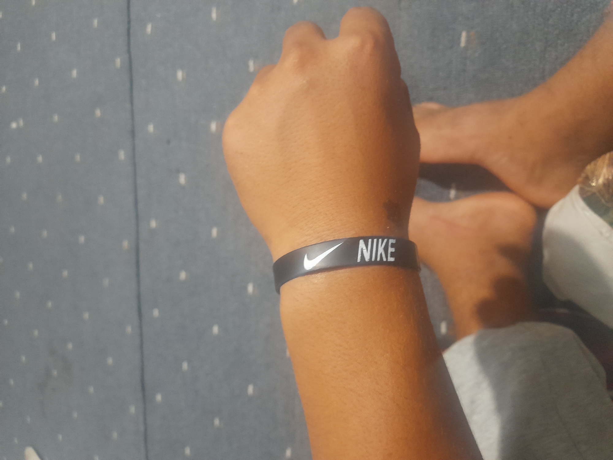 The image shows a person's arm and hand wearing a black Nike wristband. The person's legs and feet are also visible, and they are sitting on a blue carpet with a white dotted pattern. The overall scene appears to be indoors, possibly in a home setting. There is no discernible indication of the city of Кароол, Kyrgyzstan.