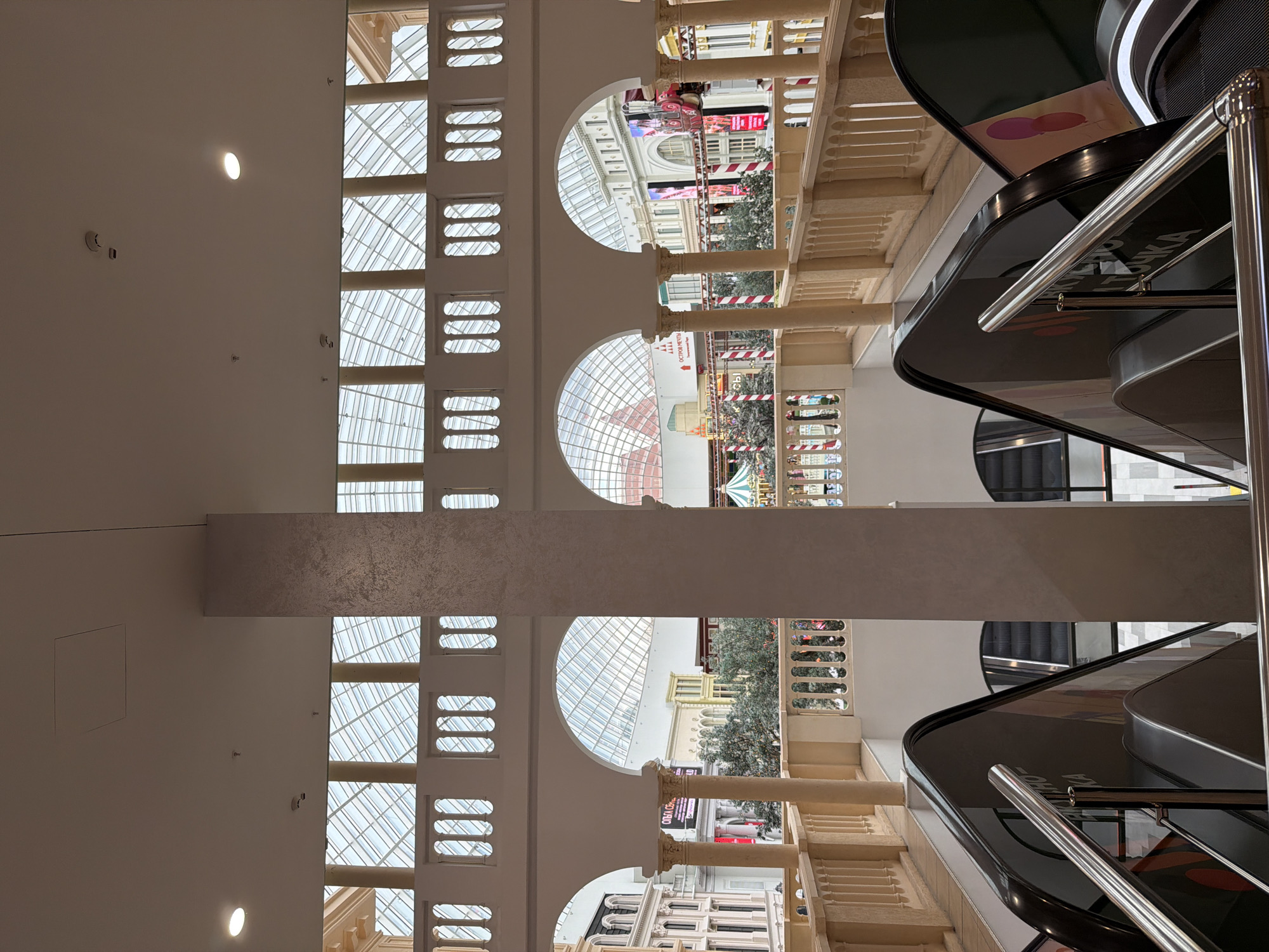 The image depicts the interior of a grand building, possibly a shopping mall or an atrium, with a focus on architectural details and escalators. The scene is bathed in natural light filtering through a large glass ceiling, suggesting it is daytime. The architecture features classic elements like arched windows, Corinthian columns, and ornamental railings, creating an elegant and spacious atmosphere.
In the foreground and right side of the image, a modern escalator with black panels and stainless steel handrails is prominent. The escalator is not in use, and no people are visible on it. Above and behind the escalator, more architectural details are visible, including stone balconies and more arched openings that look into different sections of the building.
Through the arched windows, we can see other parts of the interior, including what appears to be shops or commercial spaces with various displays and decorations. There are trees visible in some of the interior sections, adding a touch of greenery. The overall impression is one of a well-maintained, upscale commercial or public space.
On the escalator panels, some text is partially visible. One panel shows what appears to be the beginning of Cyrillic text, with the letters "ОСТРОВ" clearly legible, which translates to "Island". Another panel has a logo with two pink circles. There is also a partial display of what looks like Russian text on a red sign in the background. The image is oriented vertically.