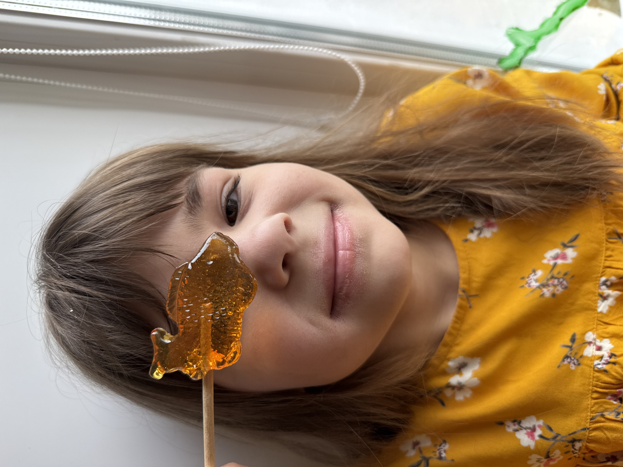 A close-up portrait shows a young girl lying down, looking towards the camera with a gentle smile. Her head is angled to the left, and her light brown hair is spread out around her. She wears a vibrant yellow dress adorned with a white and pink floral pattern.
In front of her face, she holds an amber-colored, translucent lollipop on a wooden stick. The lollipop is shaped like an animal, possibly a bird or a fish, partially obscuring her right cheek and nose. Her dark eyes and a soft smile suggest a cheerful and contented mood.
The setting appears to be indoors, brightly lit by natural light, likely from a nearby window. White horizontal window blinds are visible in the upper background, with a subtle hint of a green object further in the distance on the right. The surface beneath her is white. There is no visible text in the image. The context for the image is Rusanovo, Russia.