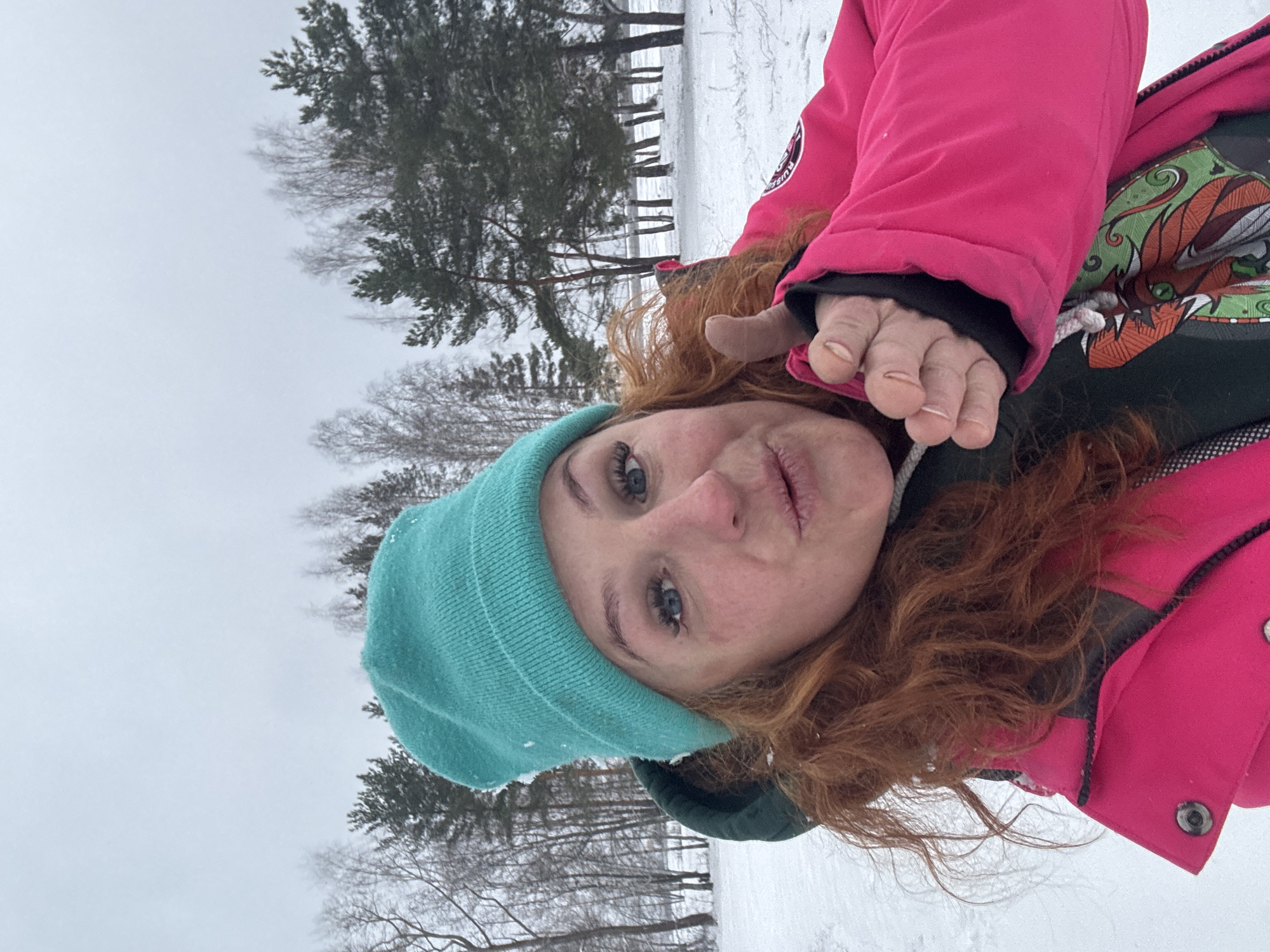 A selfie shows a woman with red hair wearing a turquoise beanie and a bright pink winter coat. She is pouting slightly at the camera and holding up her hand. The background is a snowy landscape with a line of trees. The photo was taken in Noviy, Russia, as indicated in the metadata.