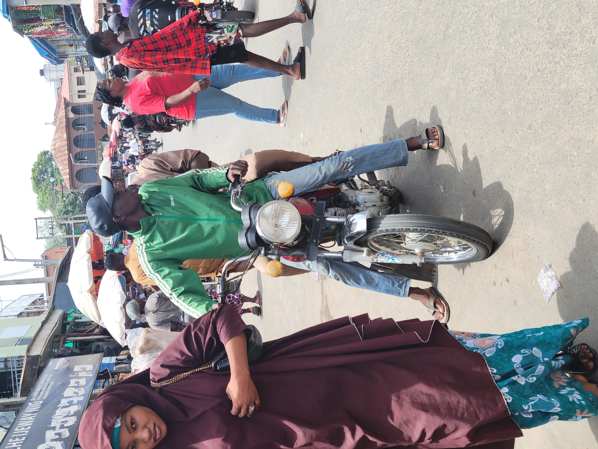 The image depicts a bustling street scene in Suleja, Nigeria. A man in a green jacket and jeans is sitting on a red and silver motorcycle in the middle of the road. A woman in a maroon hijab and a patterned dress is standing nearby. Other people are visible in the background, including individuals wearing casual clothing such as a red t-shirt and a red plaid shirt. The environment shows various shops and buildings along the street, with many stalls and umbrellas indicating market activity. The street is paved, and the overall impression is a crowded, active urban environment.