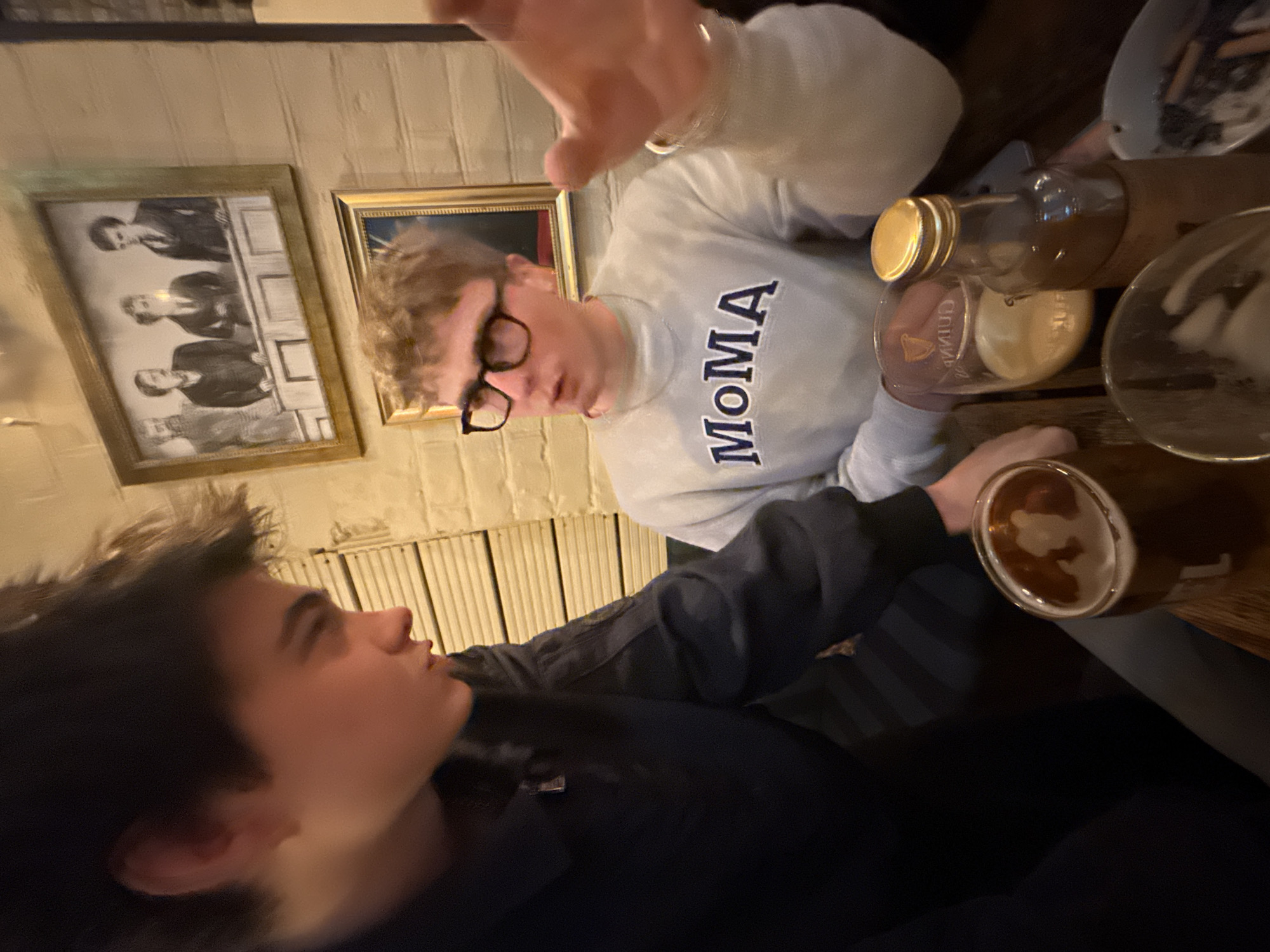 The image depicts two young men indoors, likely in a pub or bar in London, United Kingdom, given the dim lighting suggesting an evening setting. The photo is taken from a slightly elevated angle.
In the foreground on the left, a young man with dark hair is shown in profile, looking towards the right. He is wearing a dark, long-sleeved top.
To his right and slightly behind him, another young man is visible, looking in the direction of the first. He has light, possibly curly, hair and wears dark-rimmed glasses. He is dressed in a light grey turtleneck sweatshirt with the word "MOMA" emblazoned in dark blue capital letters on the chest. His right hand is raised, palm outwards, towards the top of the frame, potentially gesturing or shielding.
The two are seated at a wooden table. On the table, several items are visible: a glass containing a dark amber liquid (likely beer) in the lower right, another glass with the faint branding "Guinness," and a bottle with a gold cap. An ashtray filled with cigarette butts is also visible in the upper right quadrant of the image.
The background features a light-colored wall with a textured or panelled surface. Two framed pictures adorn the wall: one is a black and white portrait depicting multiple men, and the other, partially visible, shows a person's face. The scene portrays a casual social interaction between the two individuals.