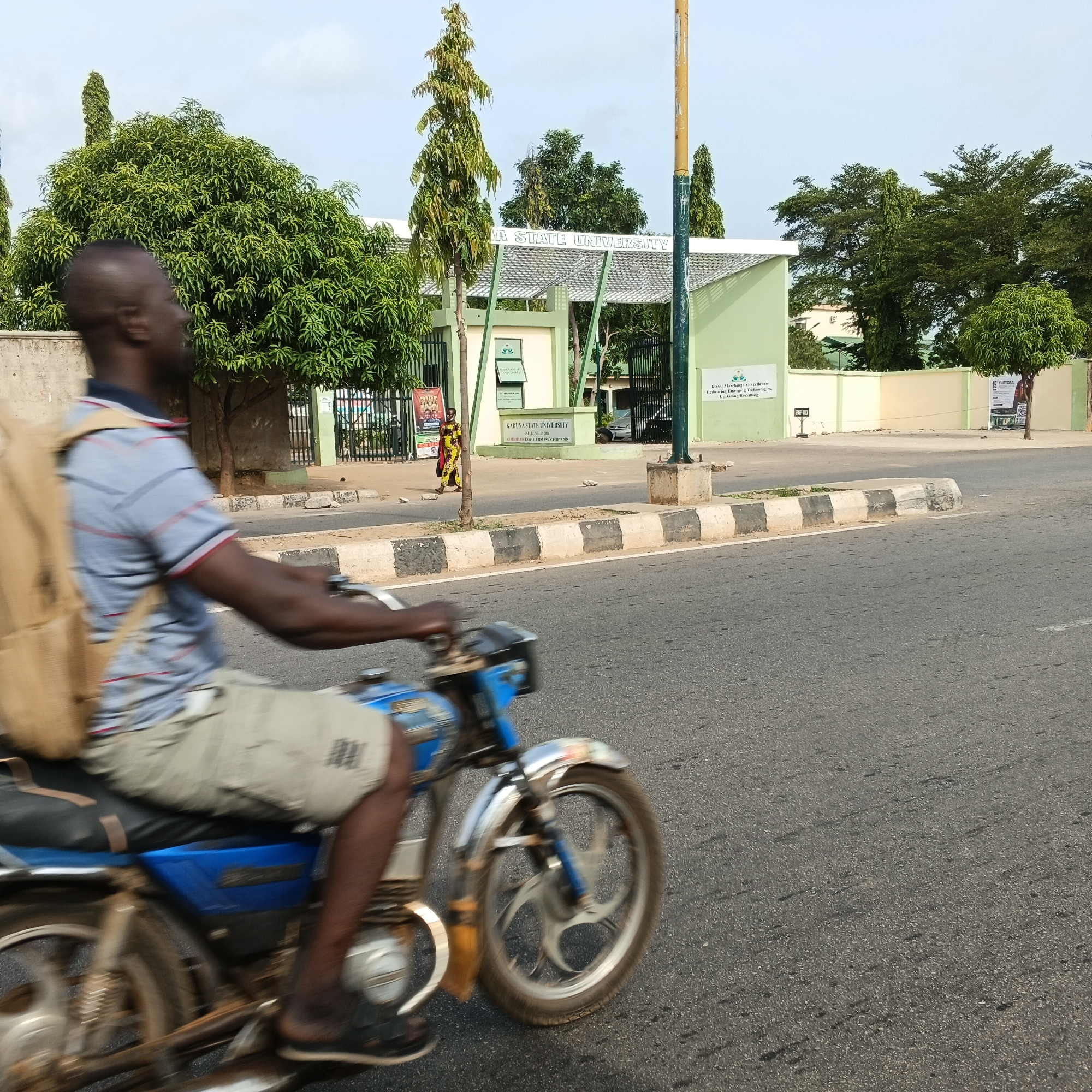 A man on a blue and silver motorcycle is captured in motion on a paved road in front of the entrance to Kaduna State University (KASU) in Kaduna, Nigeria. The man, wearing a striped polo shirt and light-colored shorts, has a light brown backpack slung over his shoulder. He is viewed from the side, his body blurred due to the motion. The motorcycle's front wheel and part of the engine are visible. In the background, the university's green and white gate is prominent, with the words "KADUNA STATE UNIVERSITY" displayed above the entrance. Several trees, including tall, slender ones and a large leafy tree, surround the university entrance, and a woman in a colorful yellow and red dress is seen walking near the gate. The road has a distinct black and white striped curb. The sky is overcast.
