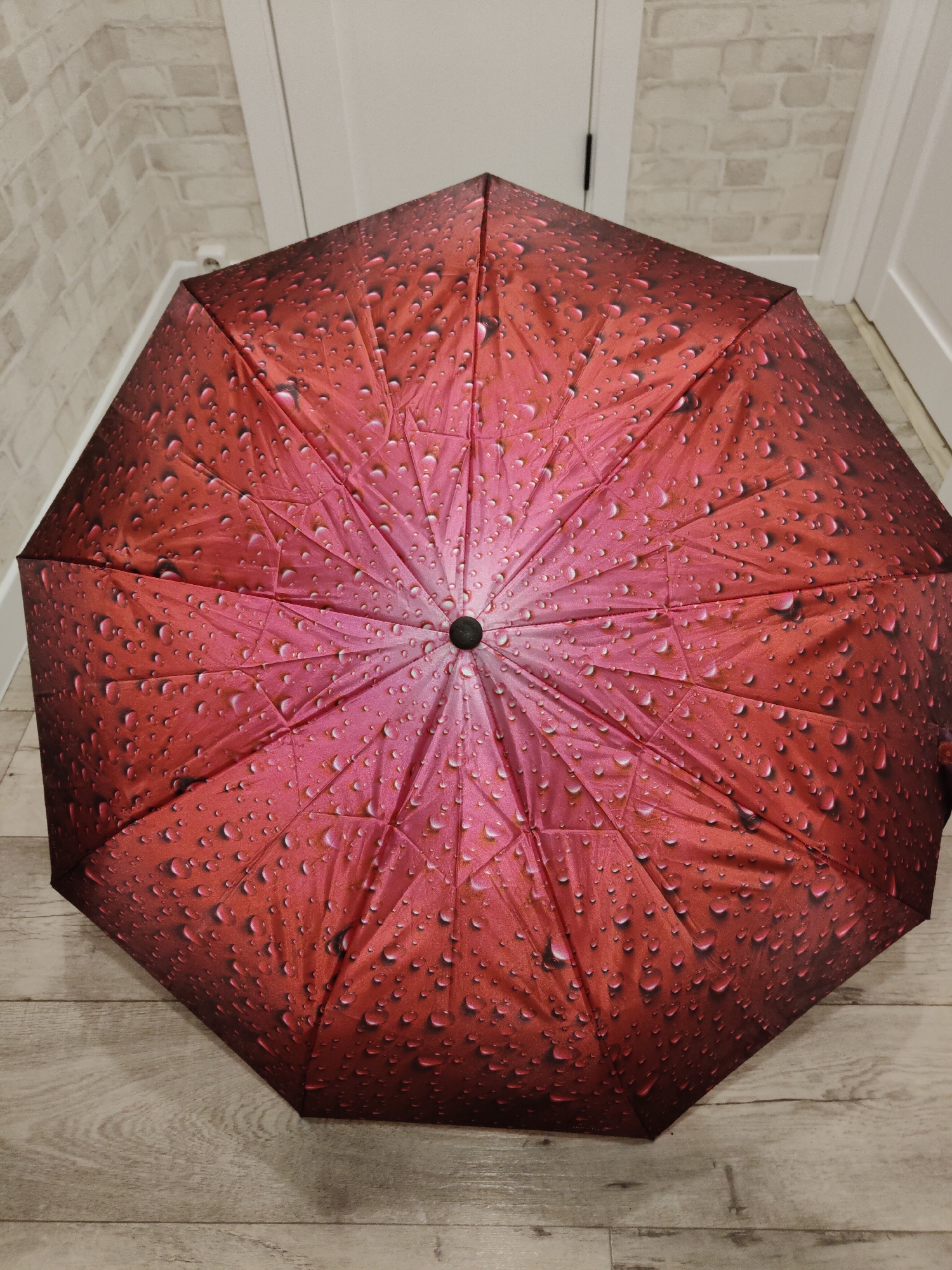 A high-angle, indoor shot presents a fully opened umbrella placed on a light-colored, wood-look floor. The umbrella has a predominantly red hue, with a gradient effect transitioning to a lighter pinkish-red towards the center. The fabric is covered with a pattern resembling water droplets. The background includes a pale brick wall on the left and a white doorframe on the right, suggesting an interior setting. The umbrella appears stationary and unoccupied.