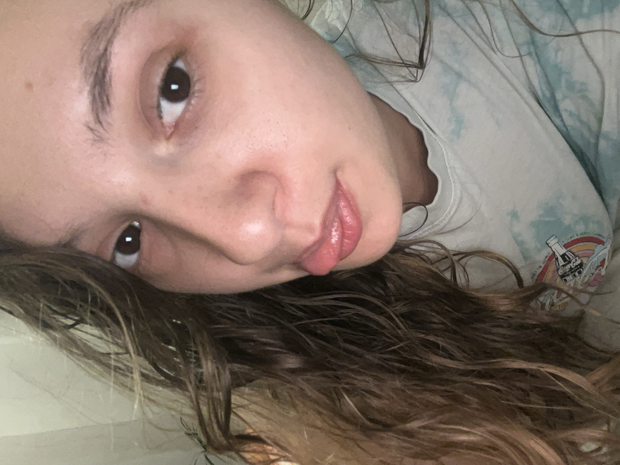 A selfie taken by a young woman with light skin, dark eyes, and brown hair. She is wearing a tie-dye t-shirt with a Coca-Cola logo and rainbow graphic. The photograph is rotated, with the woman's face angled towards the upper left of the frame. Her hair is partially obscuring the lower portion of the image. There is no indication of the location being Saarbrücken, Germany.