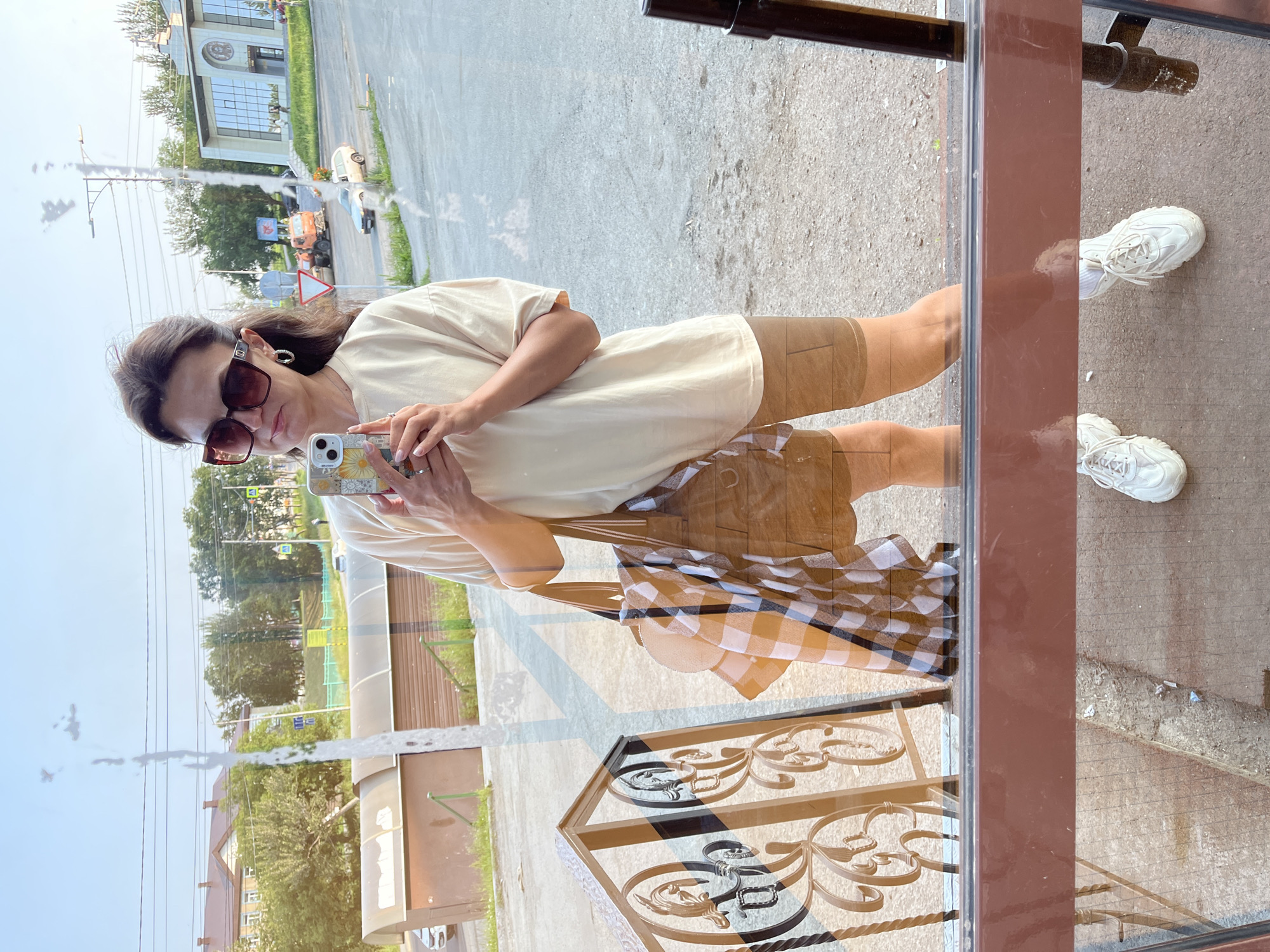 A full-body, reflected image of a woman is captured, holding a smartphone in her right hand, apparently taking a selfie. She is wearing oversized brown sunglasses, a loose-fitting, cream-colored t-shirt, and camel-colored biker shorts. A checkered orange and white scarf is draped over her left arm, which also holds a large brown tote bag. She is wearing white sneakers with white laces. The reflection shows her standing on an outdoor paved area, with a decorative metal railing to her left. The background of the reflection reveals a street scene with buildings, trees, a road with vehicles, and construction equipment, suggesting an urban environment. The image is taken from within a building, as indicated by the window frame and reflections superimposed on the scene. The context of Petropavlovsk-Kamchatsky, Russia, is not visually evident from the provided image.