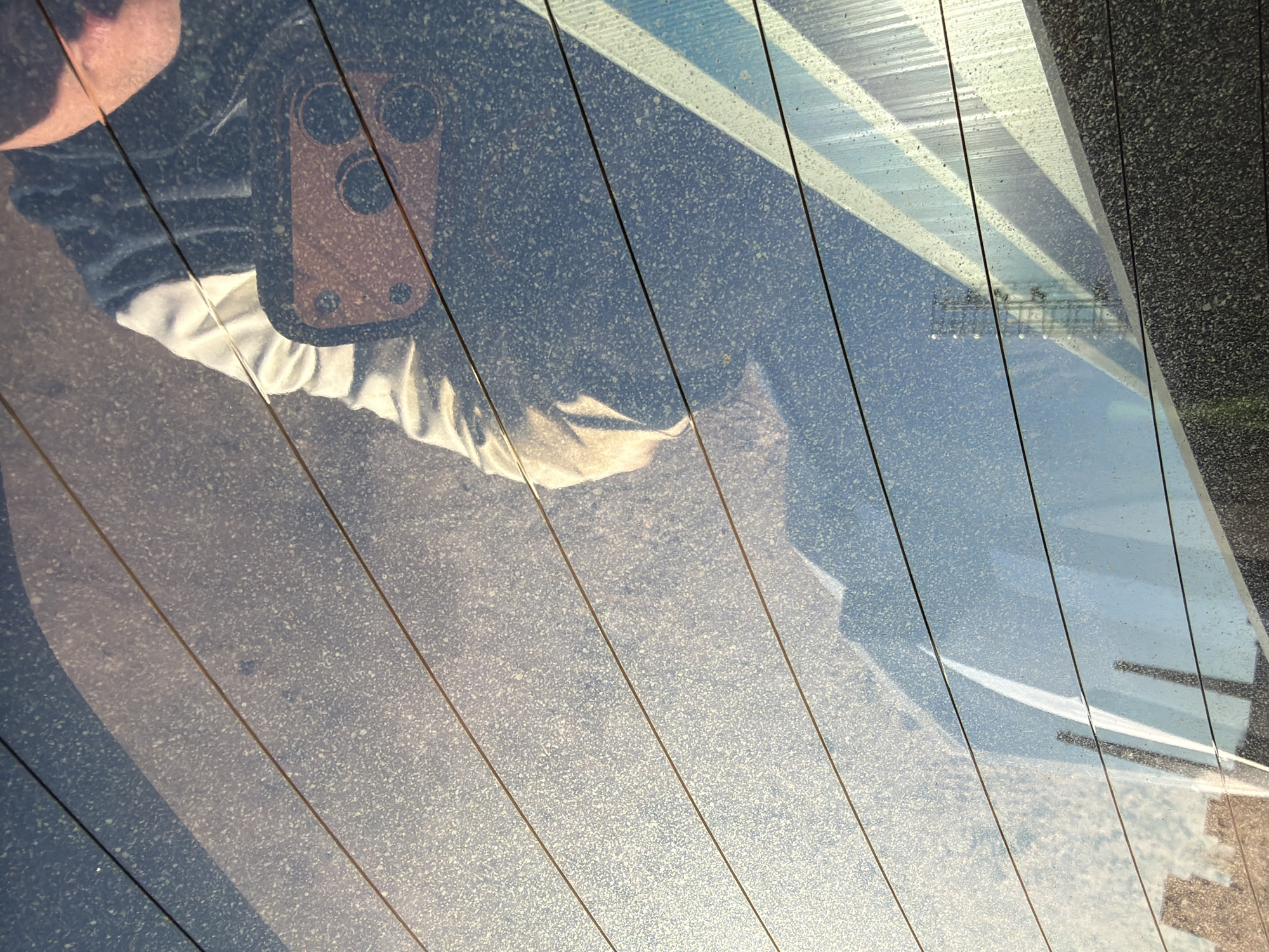 The image appears to be taken through the dirty rear window of a vehicle. The window is streaked with dust and water spots. Thin, horizontal lines are visible, likely defroster wires. The reflection of a person holding a camera is visible in the top left. The reflection suggests the person is wearing a black jacket over a light-colored shirt. Outside the window, there's a partially visible structure with blue and white stripes, possibly a roof or awning. Beyond that is what appears to be a metal structure, resembling a fence, and a gray concrete structure. It is not possible to confirm the location of the city of Chaikovsky, Russia based on visual information in this image.