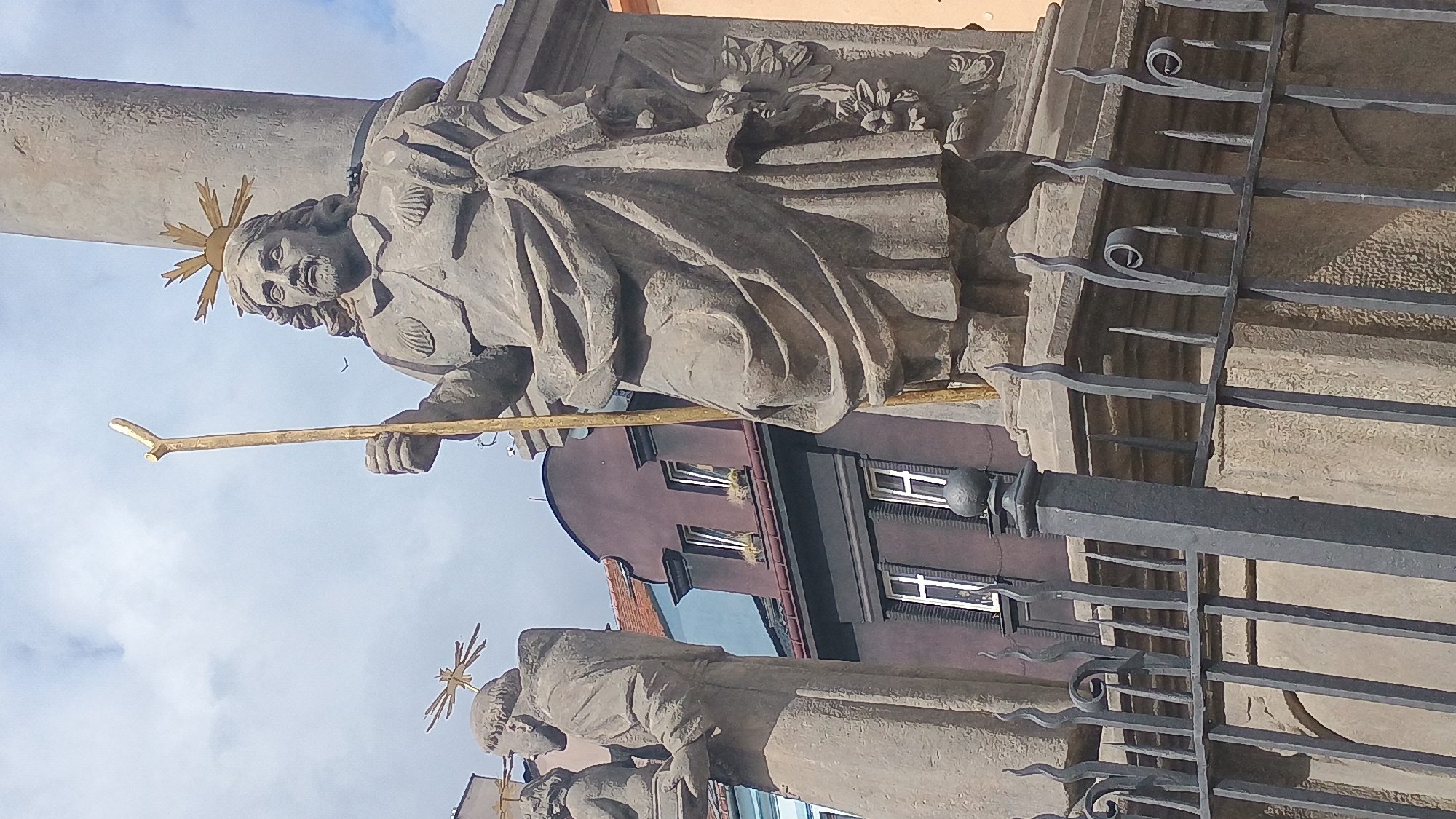 A stone statue of a bearded man holding a golden staff stands in front of a building. The man has a halo of golden rays above his head and wears a robe with two shell-shaped ornaments on his chest. Another stone statue is visible to the left, partially obscured by the main statue. In the background, a purple building with white-framed windows is visible. The statues are behind a black metal fence with decorative elements. The sky is partly cloudy. This is the statue of St. James the Greater on the square of St. James in Sokolov, Czechia.