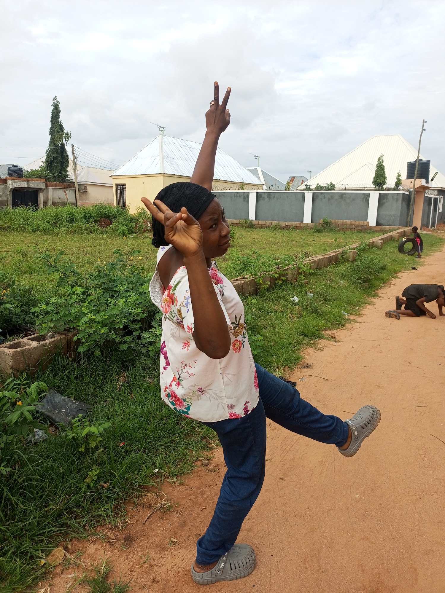 A medium shot captures a Black woman in Gombe, Nigeria, striking a pose with both hands raised in a peace sign against a backdrop of a cloudy sky and residential buildings. She wears a black headscarf, a white floral-patterned blouse, and dark blue jeans. Her right leg is extended backward, and she is wearing grey slip-on sandals. In the background, two children are visible: one is playing with a tire, and the other is kneeling on a dirt path. The environment includes overgrown green grass, a low brick wall, and a dirt road, typical of the Gombe metropolis.