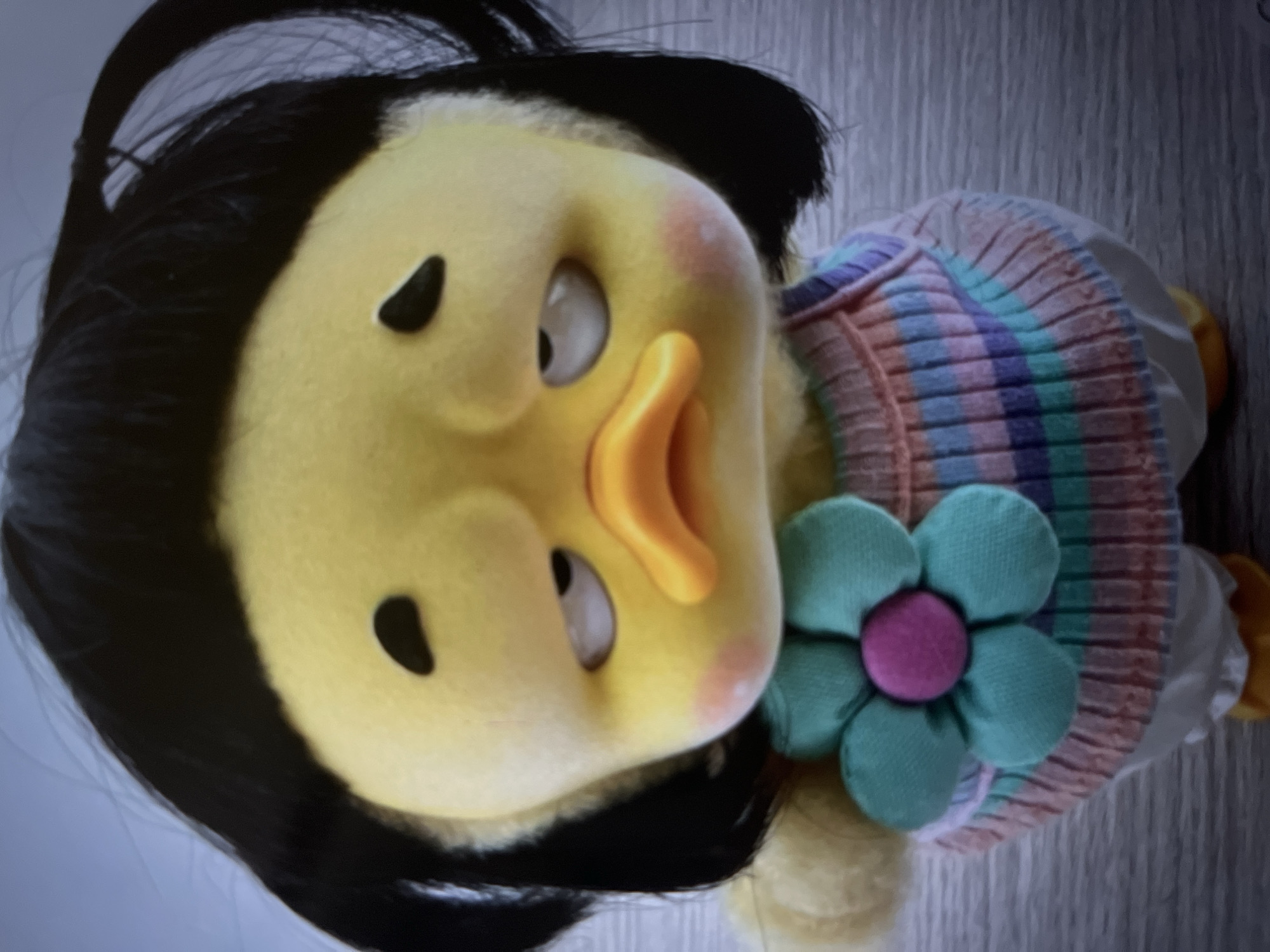 This media file is a close-up, tilted shot of a stuffed toy, likely a duck, with black hair and a yellow face. The toy is wearing a colorful, striped sweater in shades of pink, purple, blue, and green. A teal fabric flower with a pink center is attached to the sweater on the right side of the toy's chest. The toy has large, white eyes with black pupils that appear to be looking off to the side. Its beak is bright orange and protrudes from its face. The toy's cheeks have a slight pink blush. The background is a light gray wall with a subtle vertical texture. There is no visible text in the image. The lighting is soft, suggesting an indoor setting, and the overall mood is whimsical and childlike.
