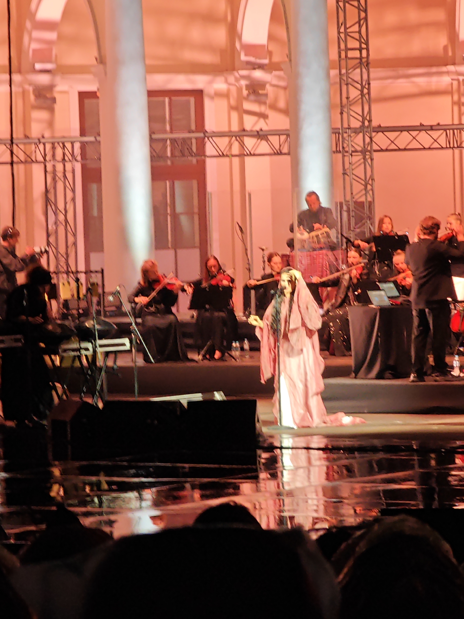The image captures a performance on a stage illuminated by warm, orange lighting. In the foreground, a female singer in a flowing, pale pink garment is kneeling and appears to be singing into a microphone. Behind her, an orchestra is performing, with several musicians playing violins and other string instruments. The musicians are dressed in dark attire. To the right of the orchestra, a man in a dark suit stands facing away from the camera, likely conducting or participating in the performance. In the background, a building with large columns and arched windows is visible. The stage floor is reflective, showing the lights and performers. The audience is mostly obscured in the foreground, appearing as dark silhouettes. The overall scene suggests an indoor concert or theatrical event. While the image itself does not provide a specific location within Moscow, the architectural style of the building in the background, with its grand columns and classical design, is reminiscent of many cultural institutions in Moscow, such as theaters or concert halls.