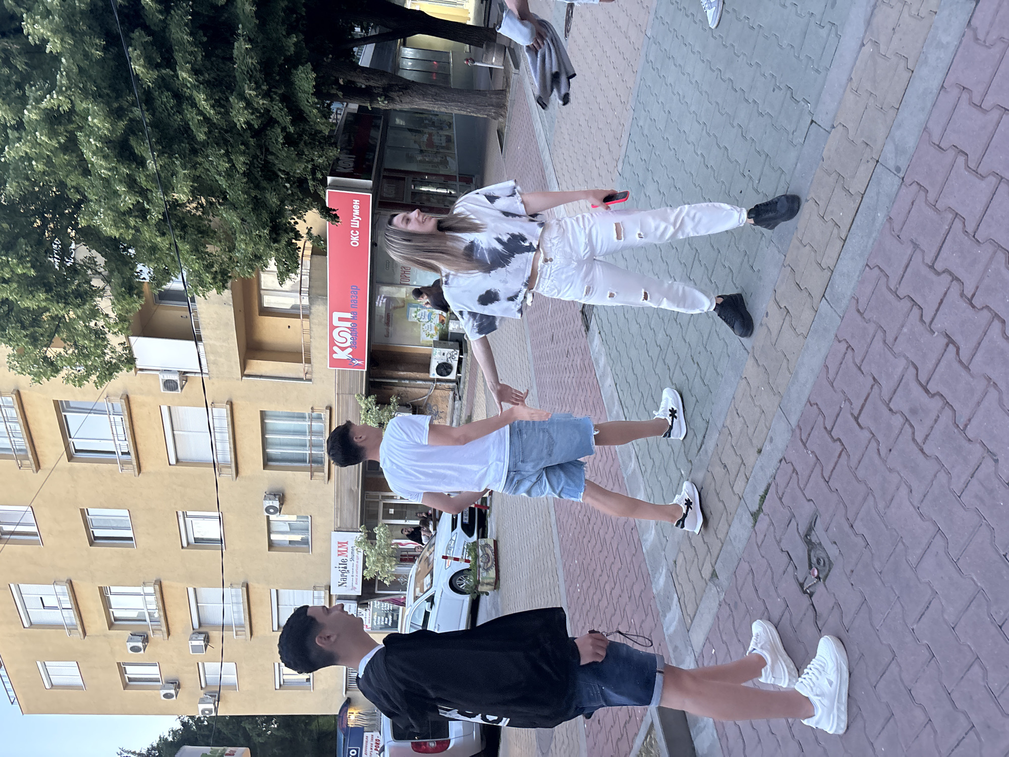 The image depicts an outdoor scene on a paved sidewalk in the city of Shumen, Bulgaria. In the foreground, three young people are interacting. A male on the left, wearing a black hoodie and denim shorts, stands looking to his right. A second male, in a white t-shirt and denim shorts, is walking away from the viewer and looking back at the female. The female, dressed in a tie-dye cropped t-shirt and ripped white jeans, is facing the male in white and appears to be gesturing or reaching out towards him. She holds a pink mobile phone in her right hand.
Behind the individuals, there is a multi-story building with yellow facade. Several air conditioning units are visible on the building's exterior. A white car is parked on the street. Signs on the ground floor of the building include "Nargile MM" and "КОП", with further text in Bulgarian. Trees with lush green foliage are present, partially obscuring the building. The ground is paved with interlocking bricks of various colors. The overall impression is a casual street scene.