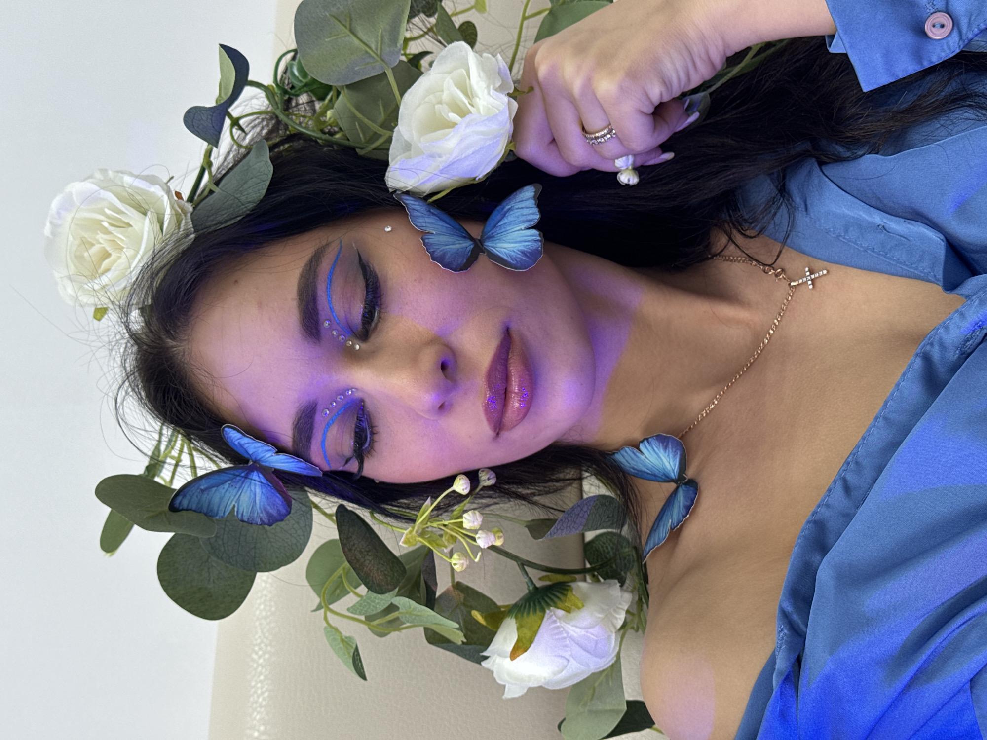 A woman with fair skin and dark hair is lying on her side, adorned with white roses, green foliage, and blue butterfly embellishments. She wears blue eyeliner with small, sparkling gems near her eyes. A gold necklace with a cross pendant rests on her chest, and a ring is visible on her hand. She is wearing a blue blouse. The background is a neutral color. There is no indication of the city of Фоки, Russia.